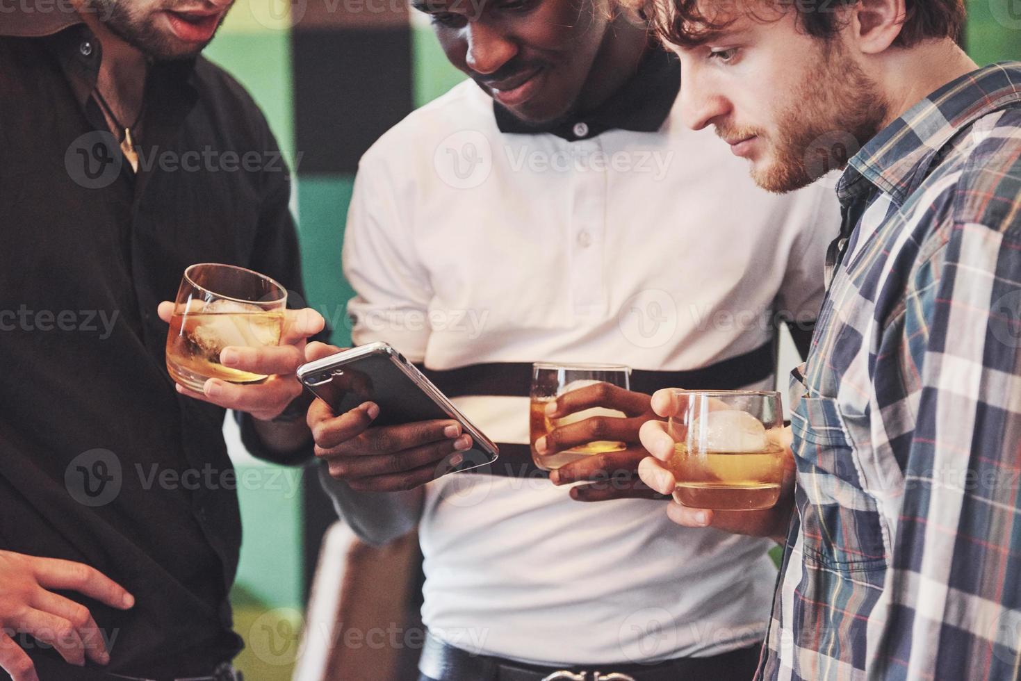 vielversprechende junge multiethnische Menschen feiern und trinken Whisky-Toast, schauen Sie sich Gadget an foto