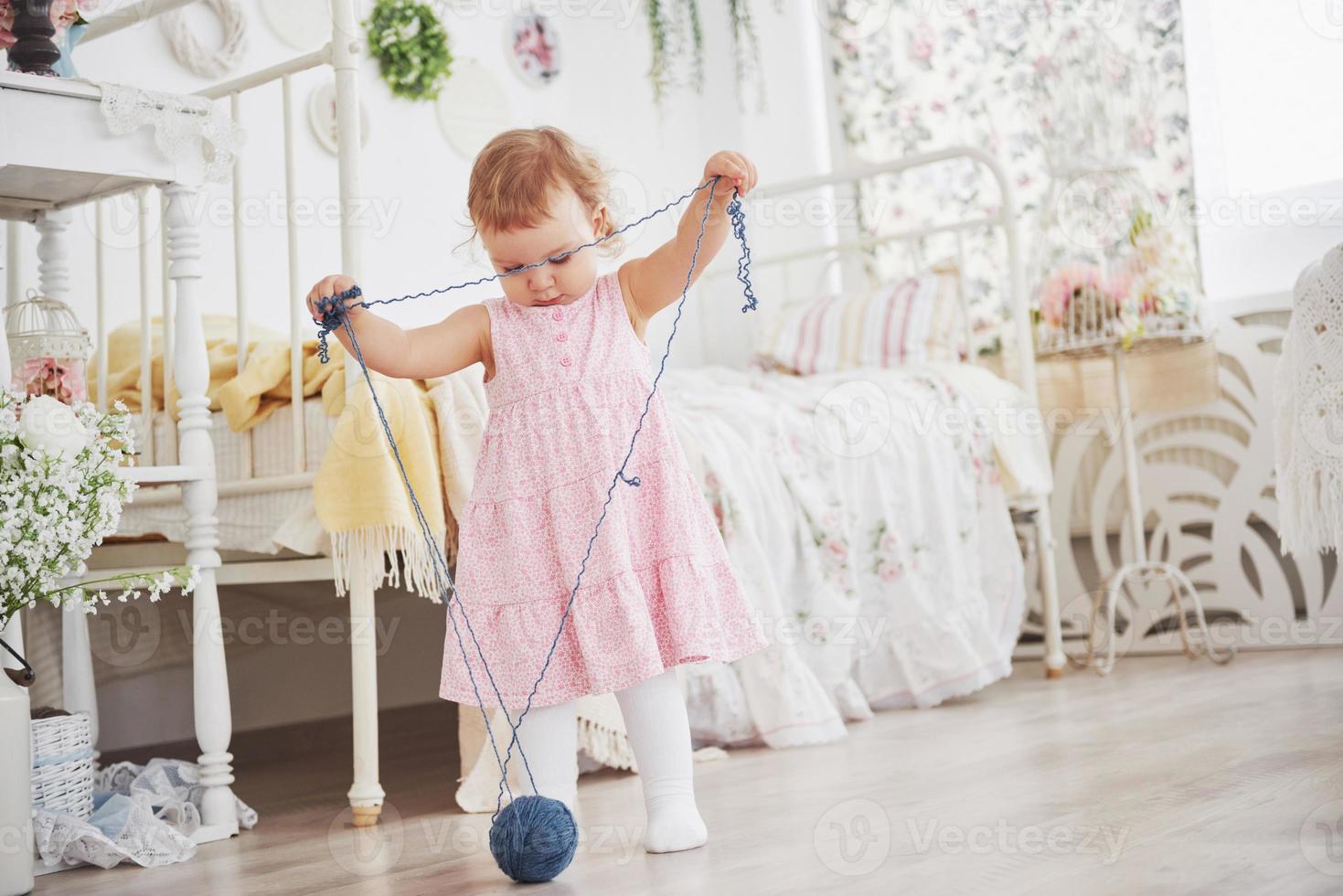 Kindheit Konzept. Babymädchen im niedlichen Kleiderspiel mit farbigem Faden. weißes Vintage-Kinderzimmer foto