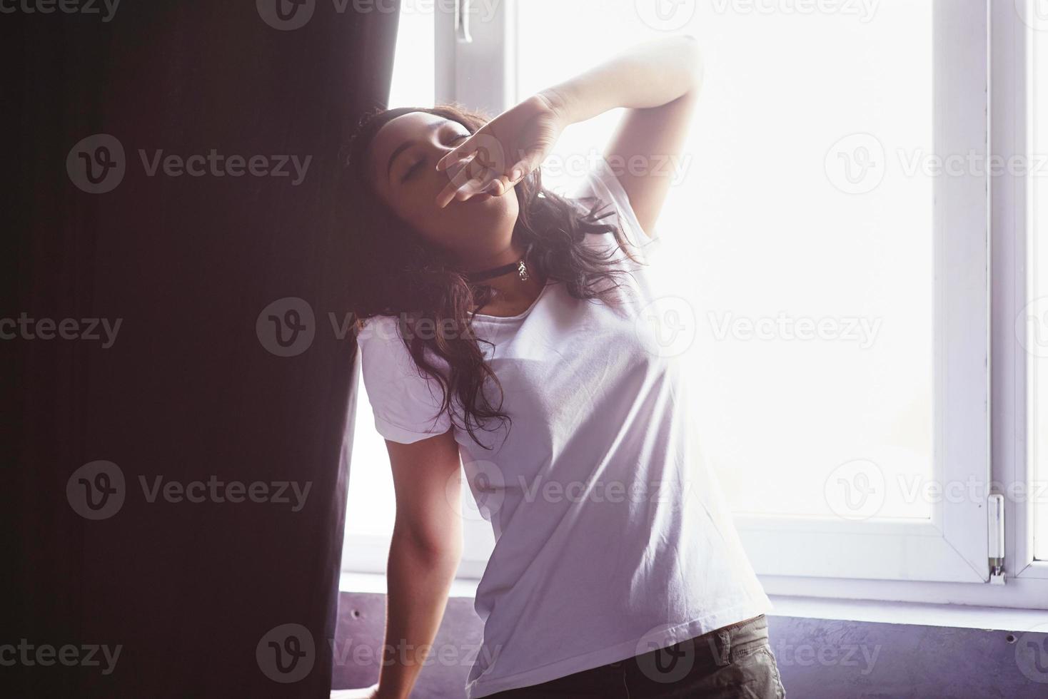 Fröhliches schönes afrikanisches Mädchen in Nachtwäsche, das sich lächelnd mit geschlossenen Augen ausdehnt, das zu Hause auf dem Bett sitzt, wachte morgens an einem sonnigen Tag auf foto