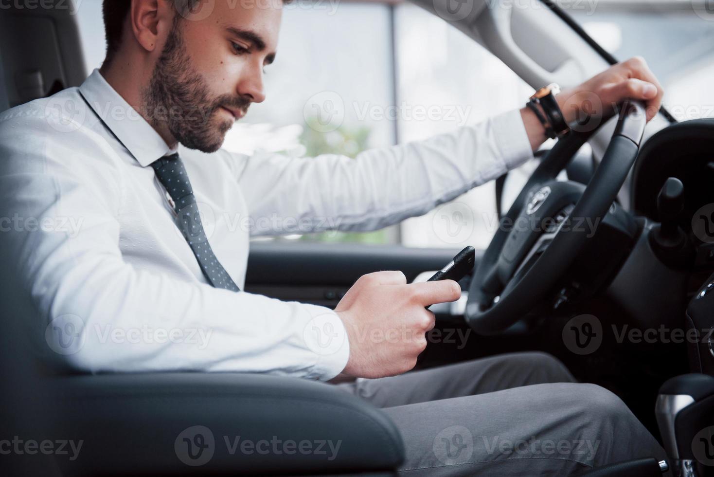 Zufriedener junger Geschäftsmann, der beim Autofahren auf das Handy schaut foto