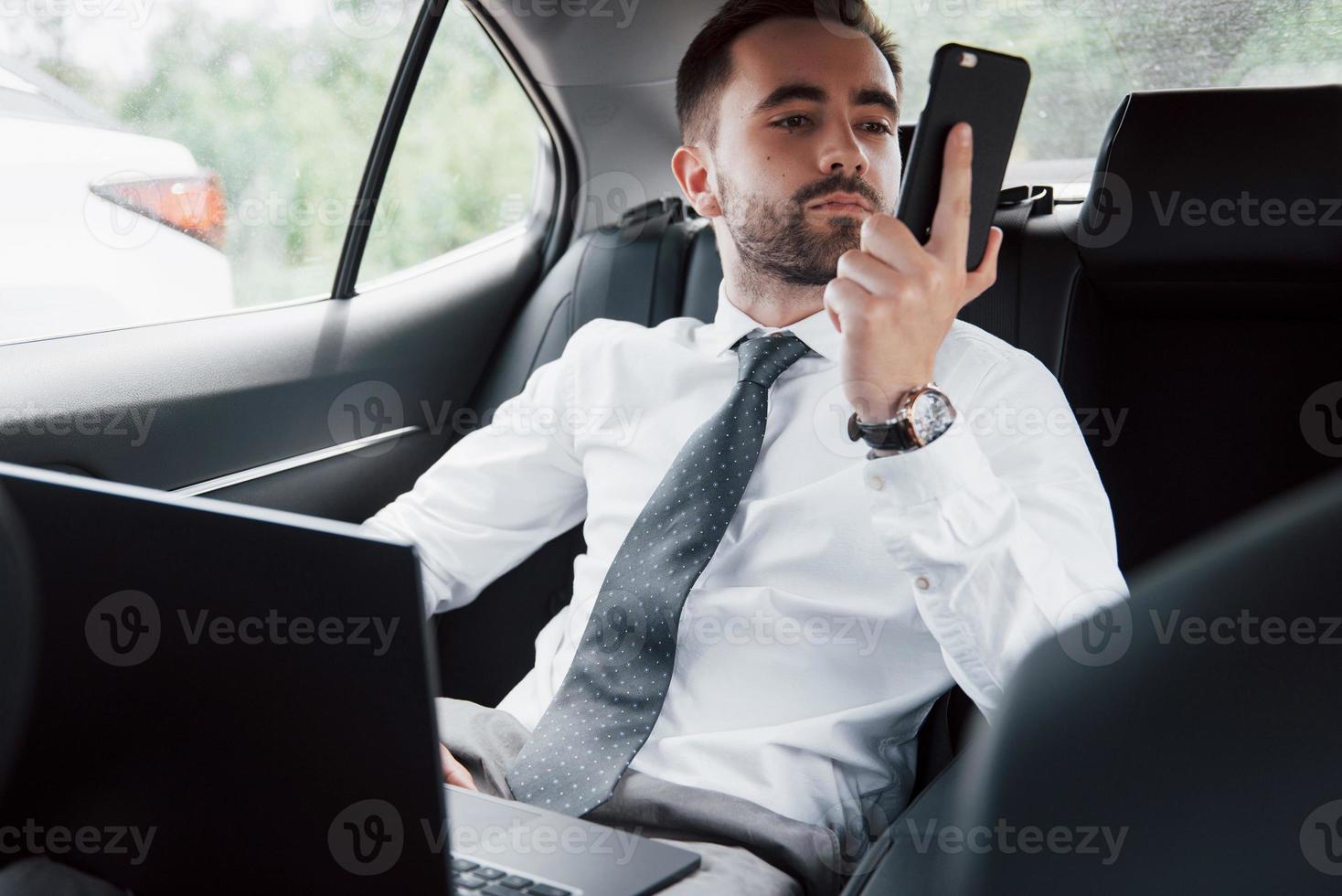 Ein junger Geschäftsmann, der am Laptop arbeitet und telefoniert, während er im Auto sitzt. arbeitet in Bewegung, schätzt seine Zeit foto