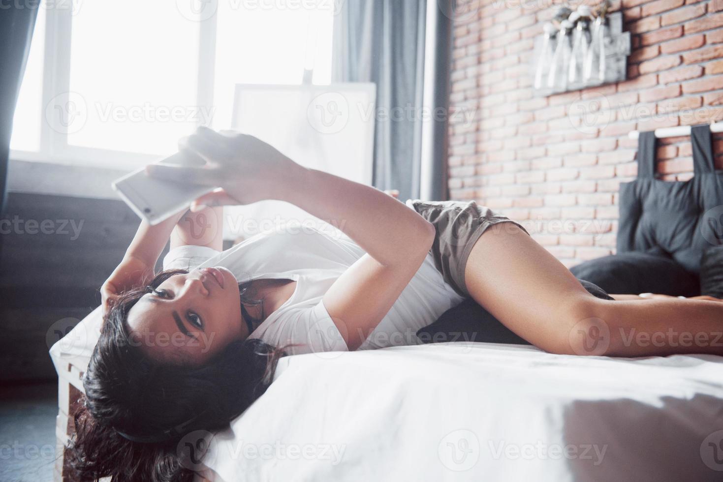 Porträt einer schönen Frau, die in ihrem Bett aufwacht und ins Telefon schaut. Überprüfen Sie soziale Netzwerke, senden Sie SMS. das Mädchen trägt ein T-Shirt foto