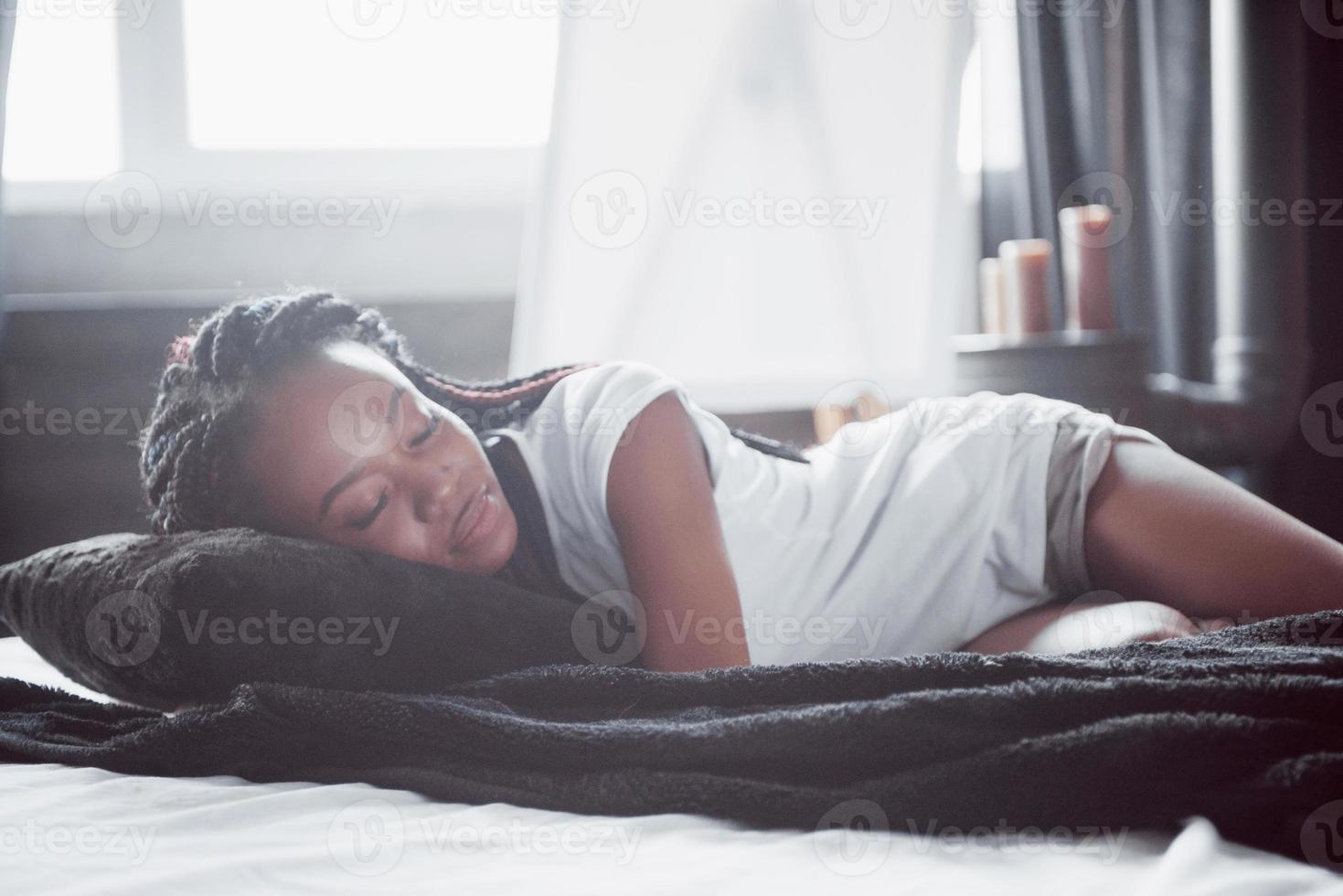 eine schöne afrikanerin liegt und schläft im bett, in einem weißen schlafzimmer. Guten Morgen, Urlaub zu Hause. das Mädchen trägt ein T-Shirt, Zöpfe auf dem Kopf foto