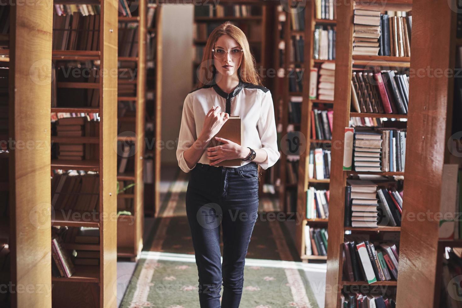 junge attraktive Studentenbibliothekarin, die ein Buch zwischen den Bücherregalen der Bibliothek liest foto