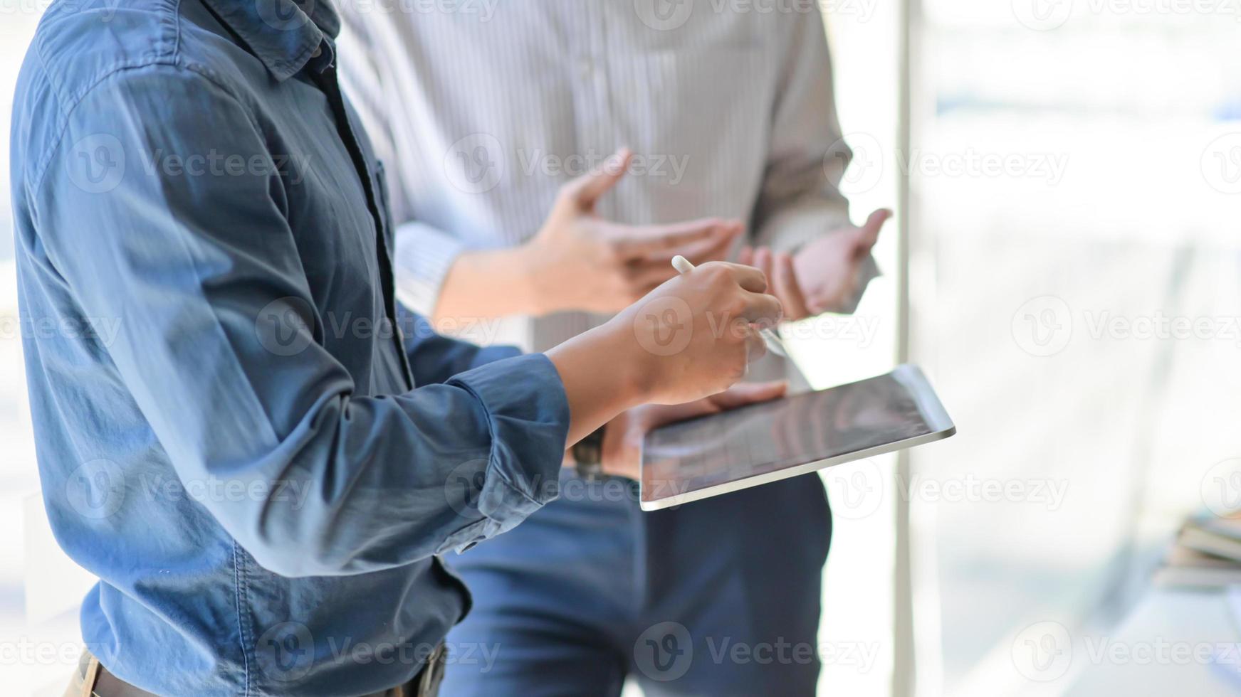 Zwei junge Geschäftsleute berieten sich über ihre Arbeit, indem sie ein Tablet benutzten, während sie am Arbeitsplatz standen, Seitenansichtsbild. foto