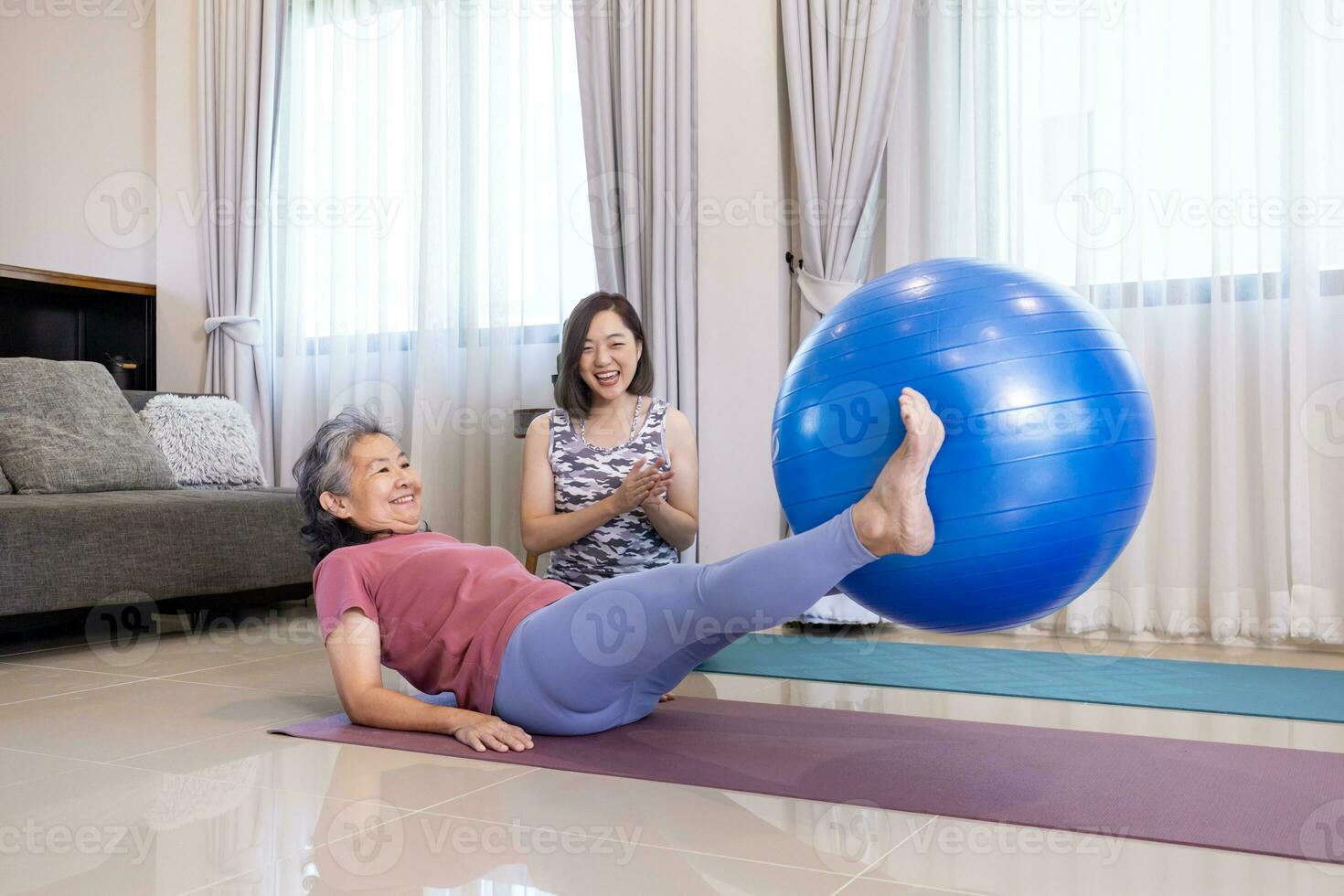 Senior asiatisch Frau ist ausüben beim Zuhause mit Yoga Ball zum Ader Körper und Abdomen Muskel Gebäude mit ihr Trainer Tochter ermutigen und Jubel zum ältere gesund Konzept foto