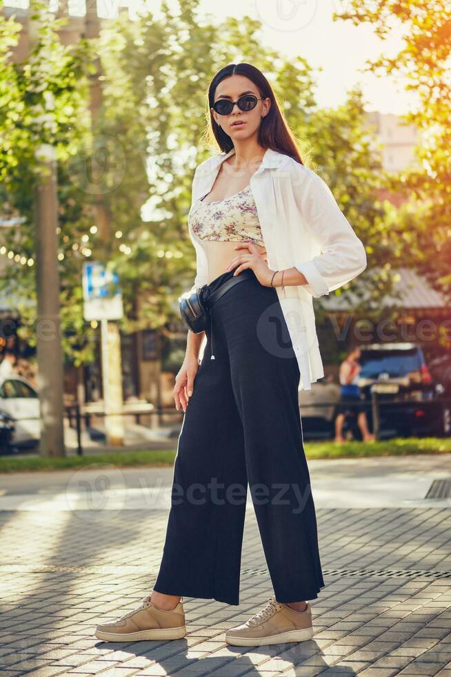 Porträt von ein Mädchen im dunkel Sonnenbrille posieren im Stadt. gekleidet im oben mit Blumen- drucken, Weiß Shirt, schwarz Hose, Taille Tasche und Turnschuhe. foto