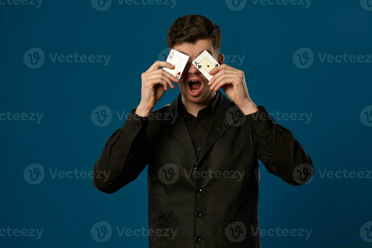 Neuling im Poker, im schwarz Weste und Shirt. halten zwei spielen Karten während posieren gegen Blau Studio Hintergrund. Glücksspiel, Kasino. Nahansicht. foto