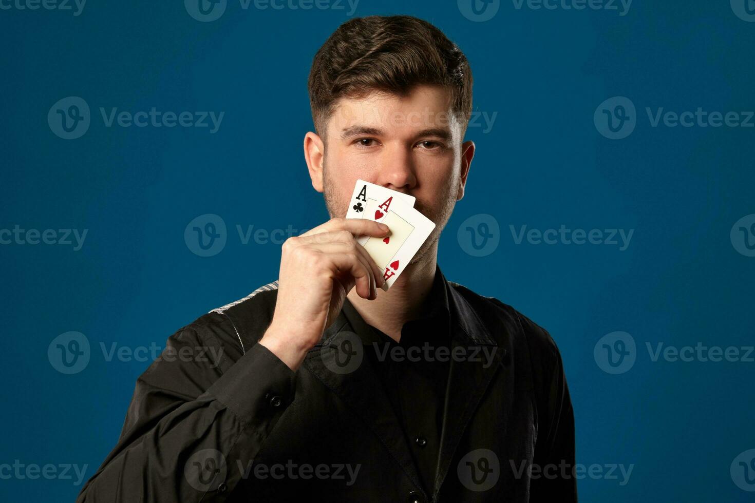 Neuling im Poker, im schwarz Weste und Shirt. halten zwei spielen Karten während posieren gegen Blau Studio Hintergrund. Glücksspiel, Kasino. Nahansicht. foto