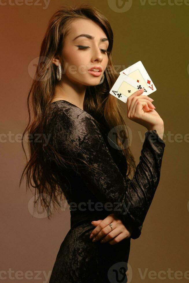 Brünette weiblich im schwarz Samt Kleid und Schmuck zeigen zwei Asse, posieren seitwärts auf bunt Studio Hintergrund. Glücksspiel, Poker, Kasino. Nahansicht. foto