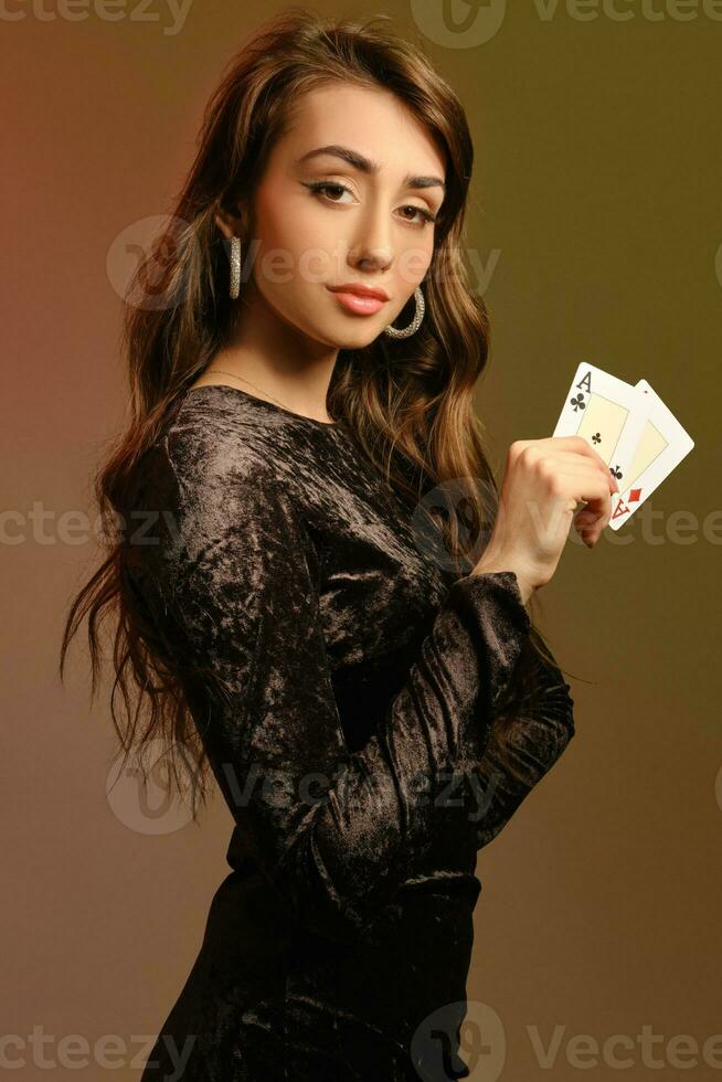 Brünette Dame im schwarz Samt Kleid und Schmuck zeigen zwei Asse, posieren seitwärts auf bunt Studio Hintergrund. Glücksspiel, Poker, Kasino. Nahansicht. foto