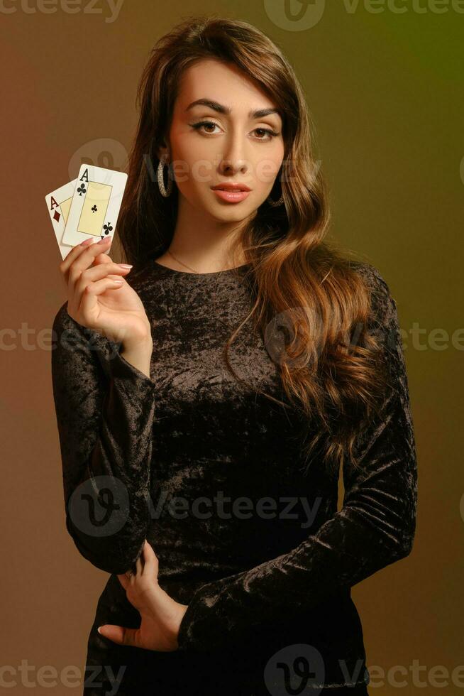 Brünette Frau im schwarz Samt Kleid und Schmuck zeigen zwei Asse, posieren gegen bunt Studio Hintergrund. Glücksspiel, Poker, Kasino. Nahansicht. foto