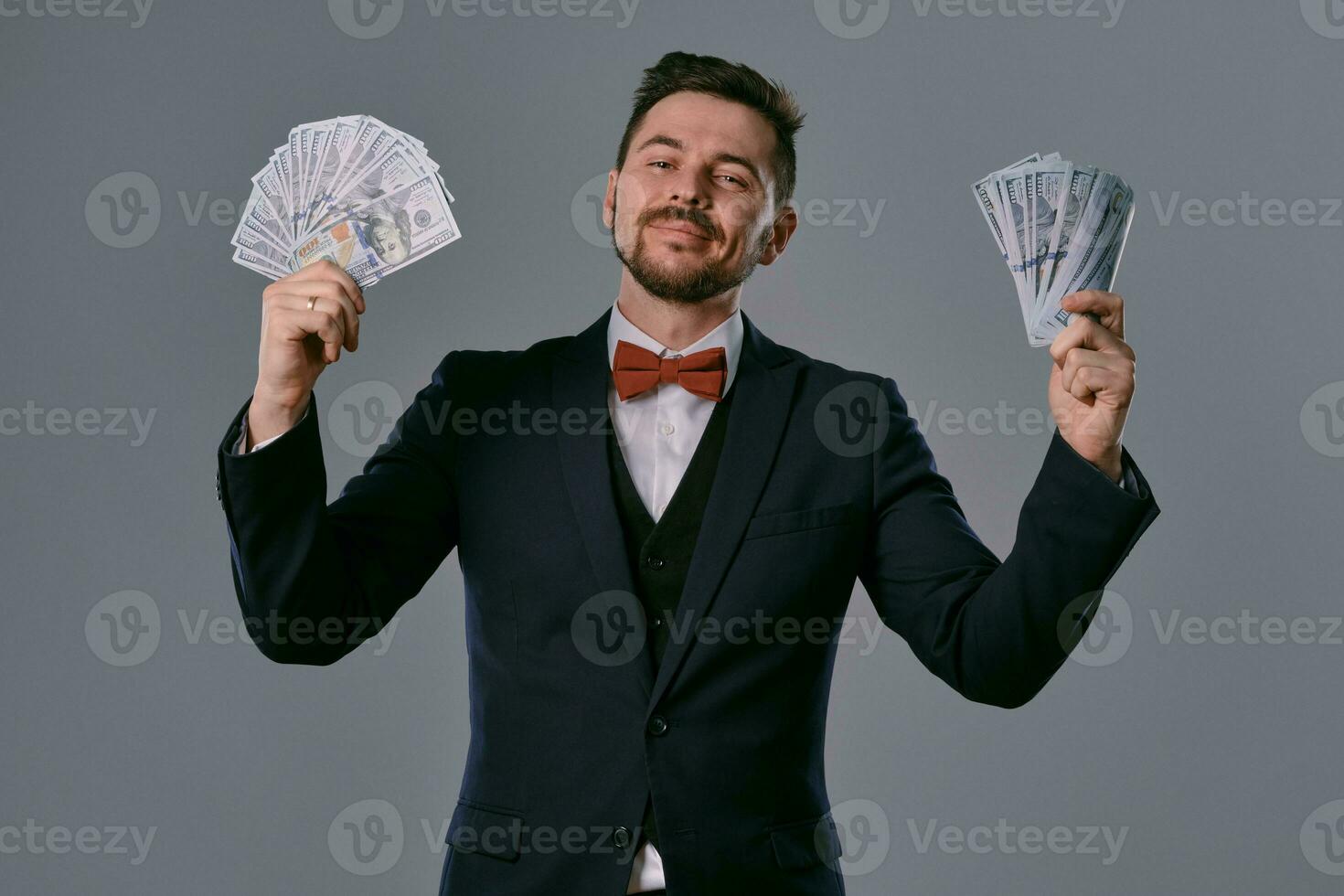 Mann im schwarz passen und rot Krawatte ist zeigen zwei Fans von hundert Dollar Rechnungen, posieren auf grau Studio Hintergrund. Glücksspiel, Poker, Kasino. Nahansicht. foto