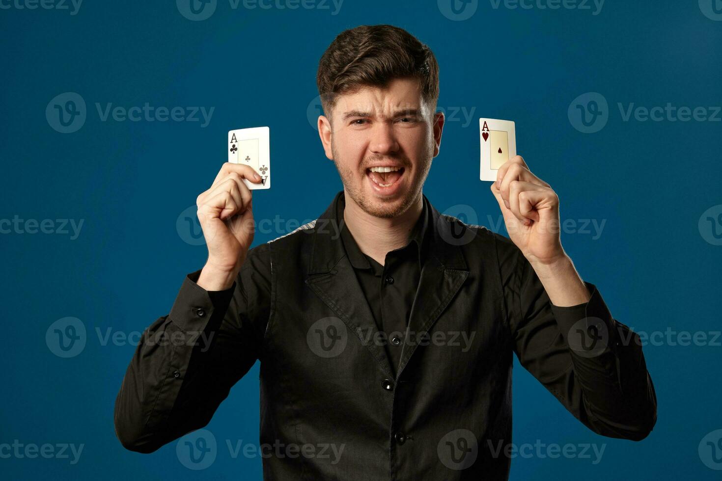Neuling im Poker, im schwarz Weste und Shirt. halten zwei spielen Karten während posieren gegen Blau Studio Hintergrund. Glücksspiel, Kasino. Nahansicht. foto