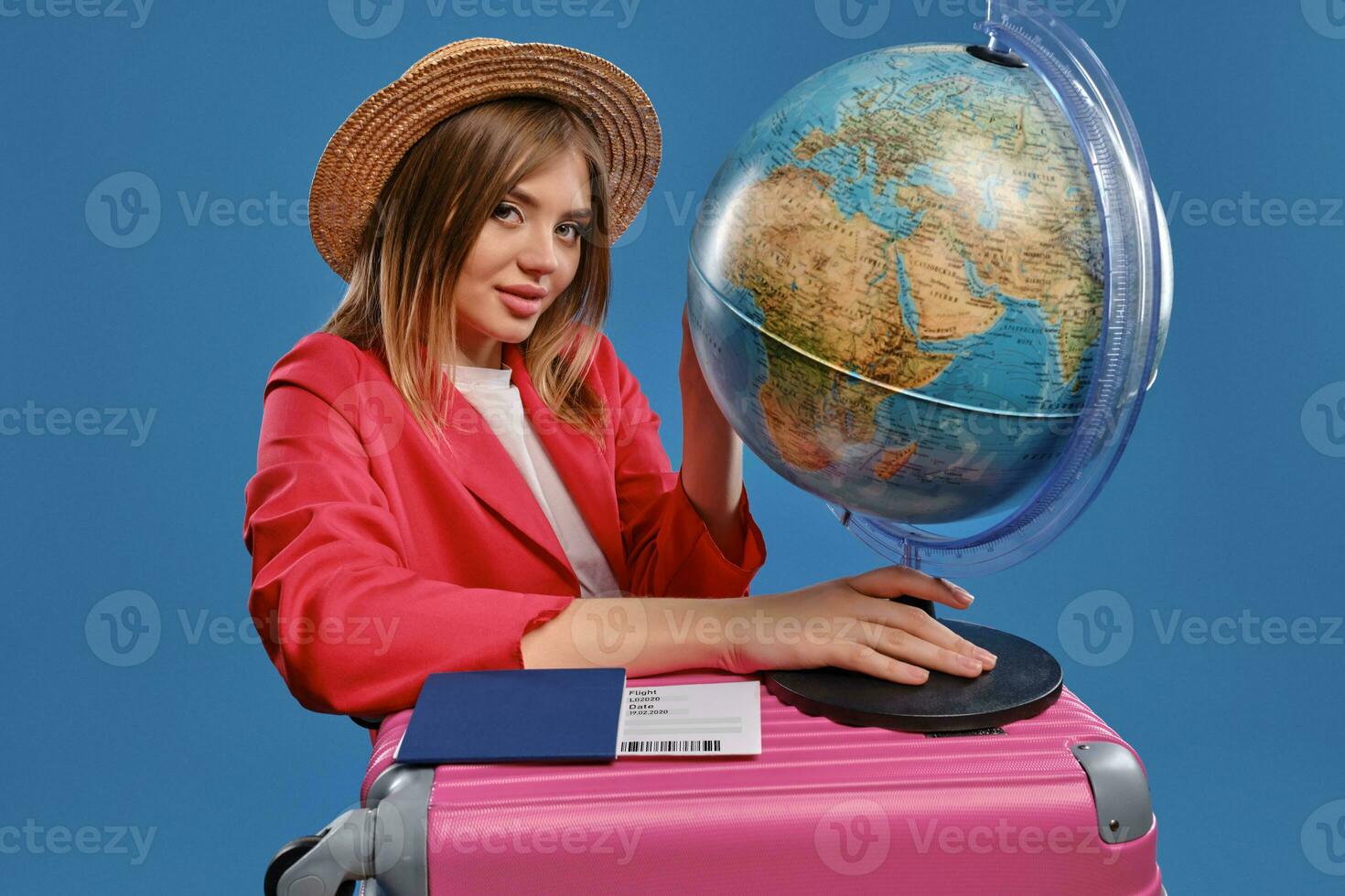 blond Dame im Stroh Hut, Weiß Bluse, rot Jacke. halten Globus Stehen auf Rosa Koffer, Reisepass und Fahrkarte nahe, posieren auf Blau Hintergrund foto