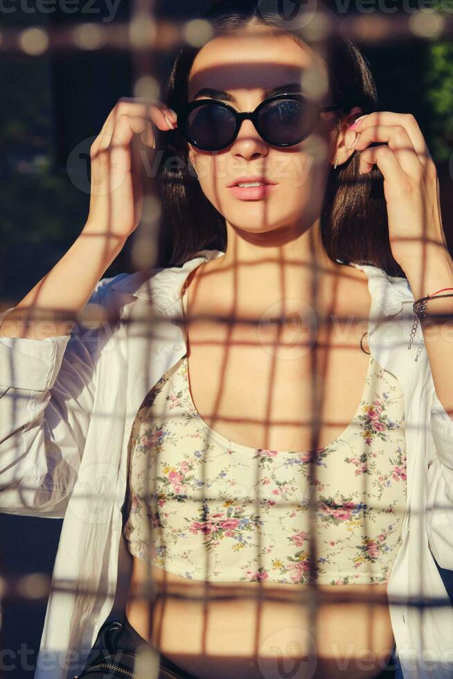 Porträt von Mädchen im Sonnenbrille posieren im Stadt hinter ein gitterförmig Zaun. gekleidet im oben mit Blumen- drucken, Weiß Shirt, schwarz Hose, Taille Tasche. foto