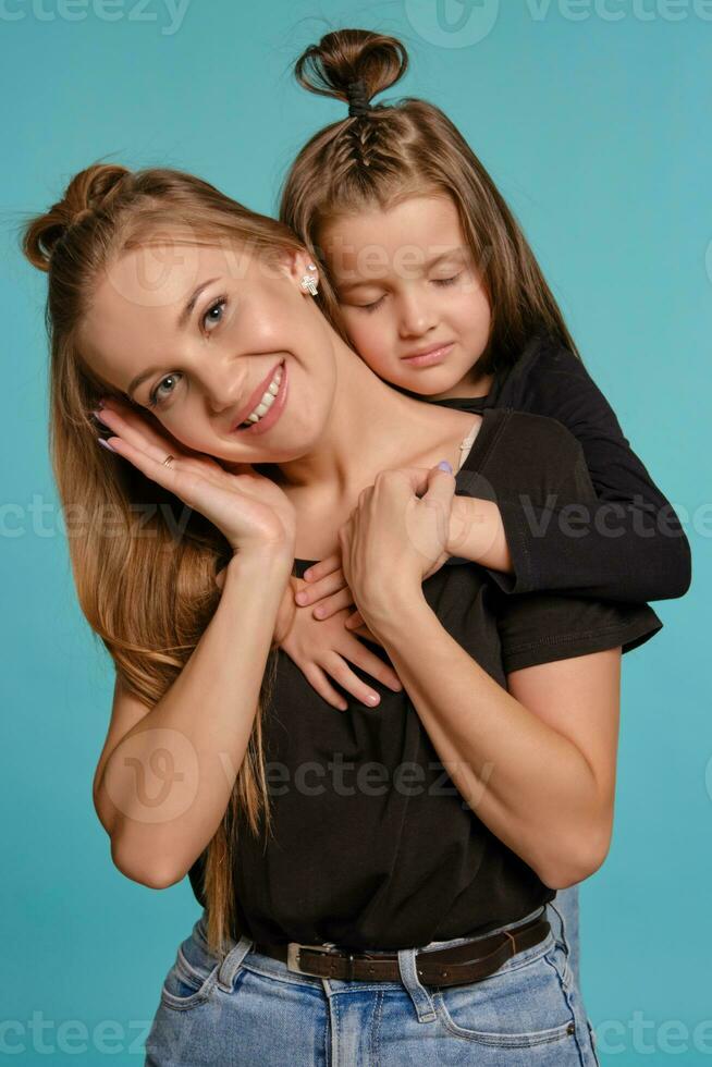 Mama und Tochter mit ein komisch Frisuren, gekleidet im schwarz Hemden und Blau Denim Jeans sind posieren gegen ein Blau Studio Hintergrund. Nahansicht Schuss. foto