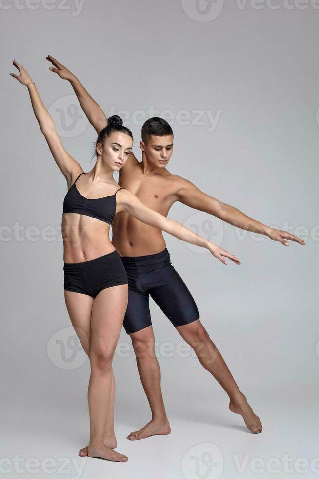 das Paar von ein jung modern Ballett Tänzer im schwarz Anzüge sind posieren Über ein grau Studio Hintergrund. foto