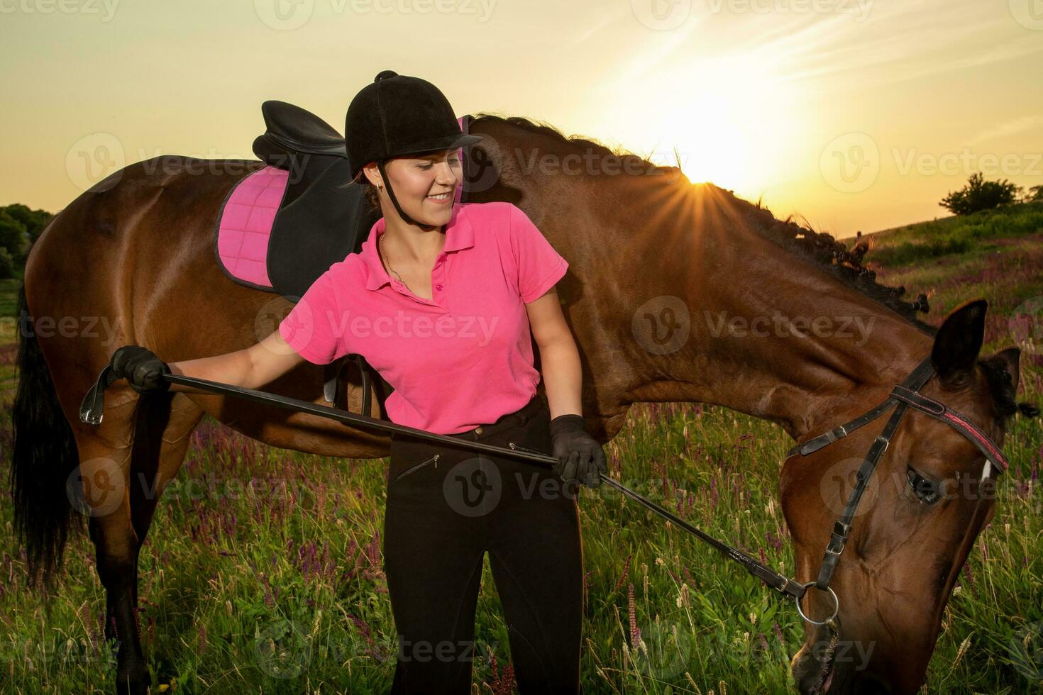 schön lächelnd Mädchen Jockey Stand Nächster zu ihr braun Pferd tragen Besondere Uniform auf ein Himmel und Grün Feld Hintergrund auf ein Sonnenuntergang. foto