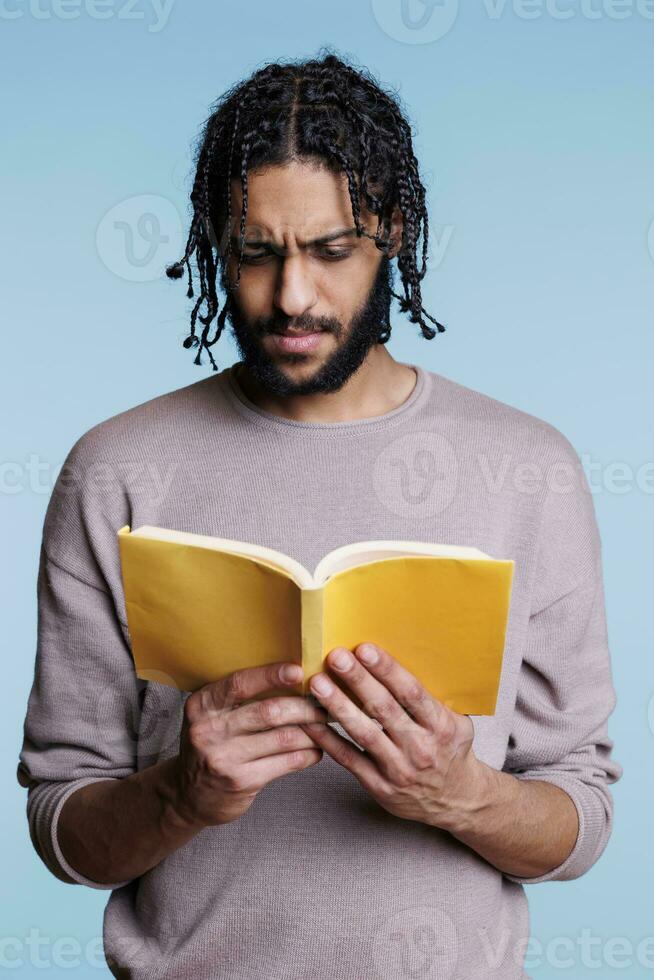 nachdenklich konzentriert arabisch Schüler lesen Roman Buch mit Gelb Abdeckung. konzentriert nachdenklich jung Erwachsene Mann studieren Literatur während halten weiche Abdeckung Taschenbuch öffnen Lehrbuch foto