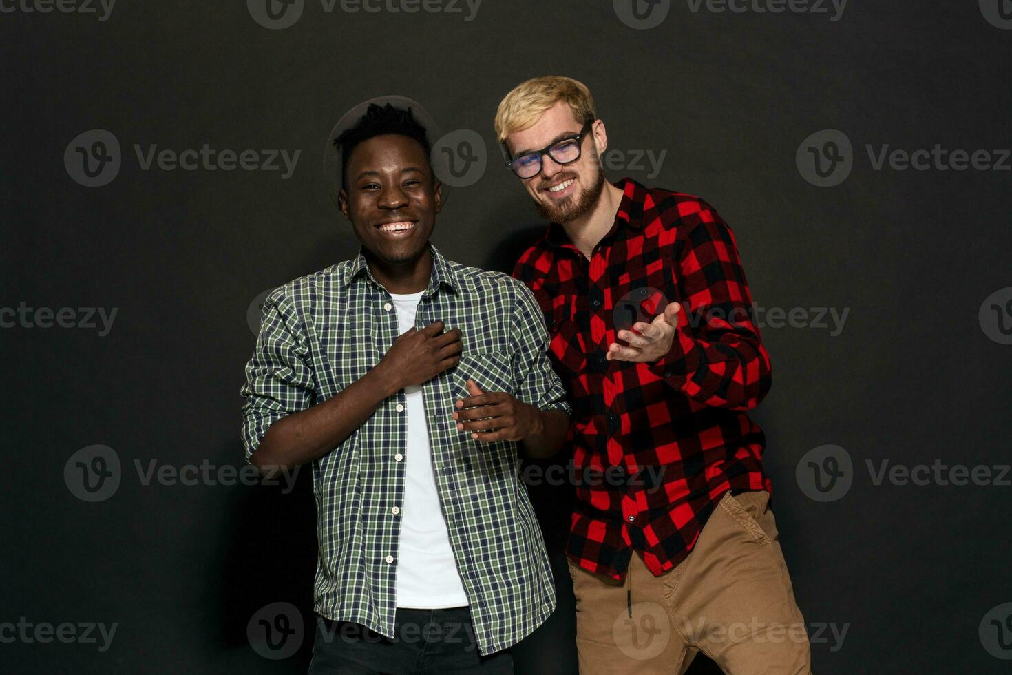 Studio Schuss von zwei stilvoll jung Männer haben Spaß. gut aussehend bärtig Hipster im ein Hemd im ein Käfig Stehen Nächster zu seine Afroamerikaner Freund im Hut gegen ein dunkel Hintergrund. foto