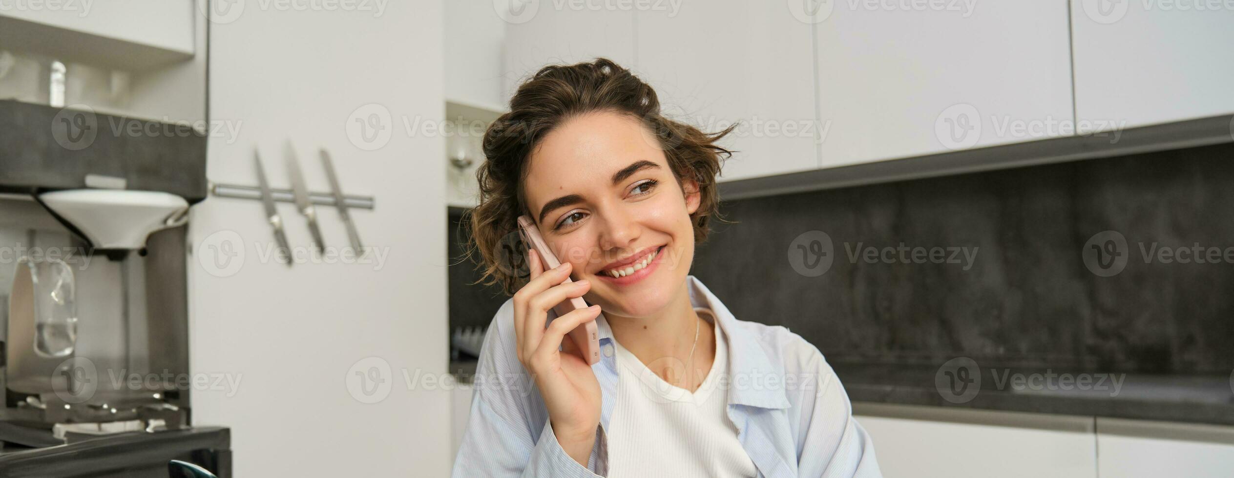 Porträt von jung Frau, 25 Jahre alt, halten Smartphone, reden auf Handy, Mobiltelefon Telefon, Sitzung im Küche beim Zuhause foto