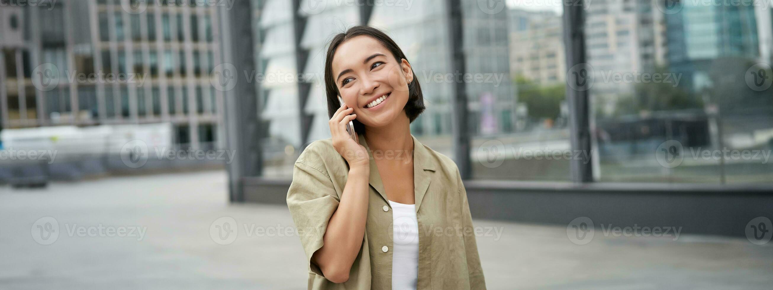lächelnd jung Koreanisch Mädchen reden auf Handy, Mobiltelefon Telefon und Gehen im Stadt. glücklich Frau posieren auf Straße mit Smartphone foto
