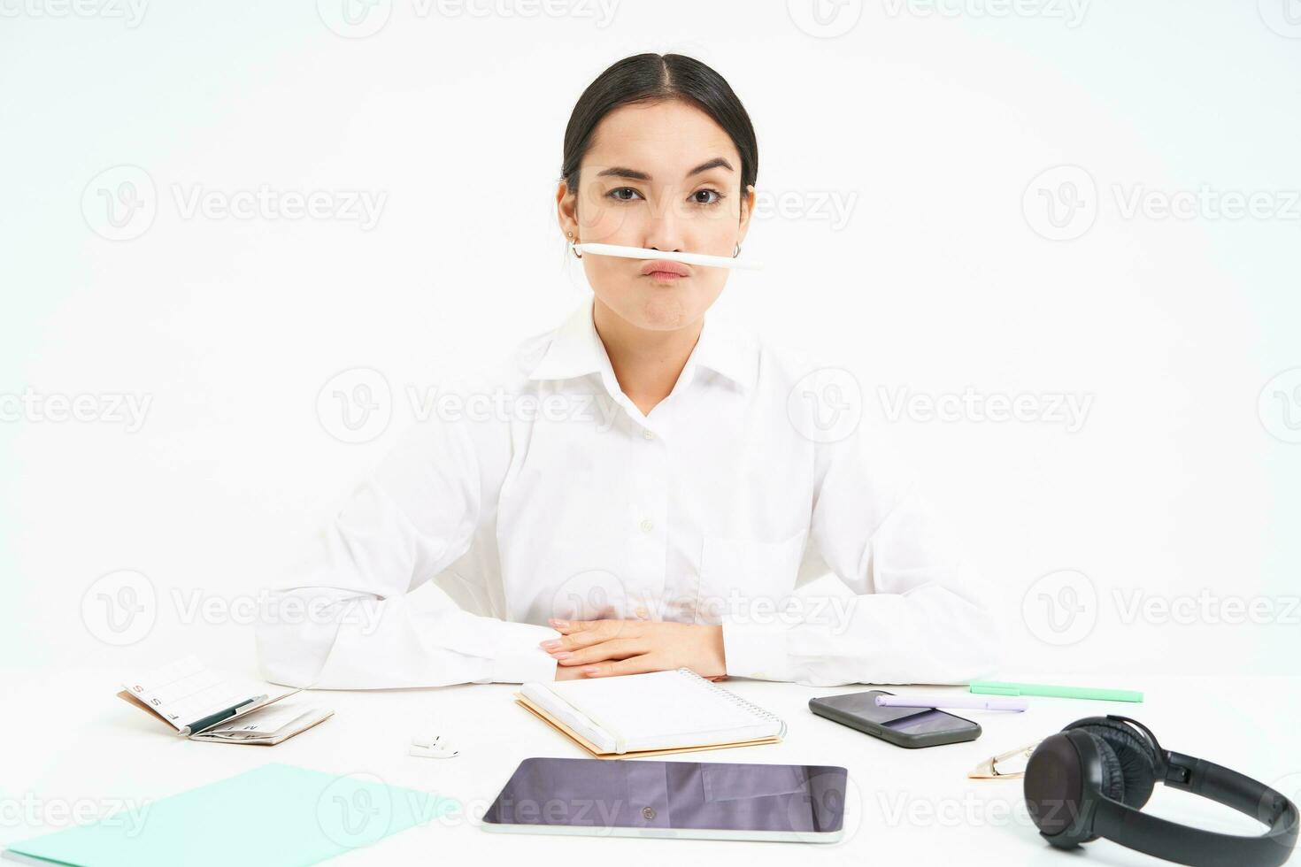 Geschäft und Büro arbeiten. jung Frau Sitzung beim Arbeitsplatz, täuschen um, hält Stift mit Lippen und zeigt an komisch Gesichter, Weiß Hintergrund foto