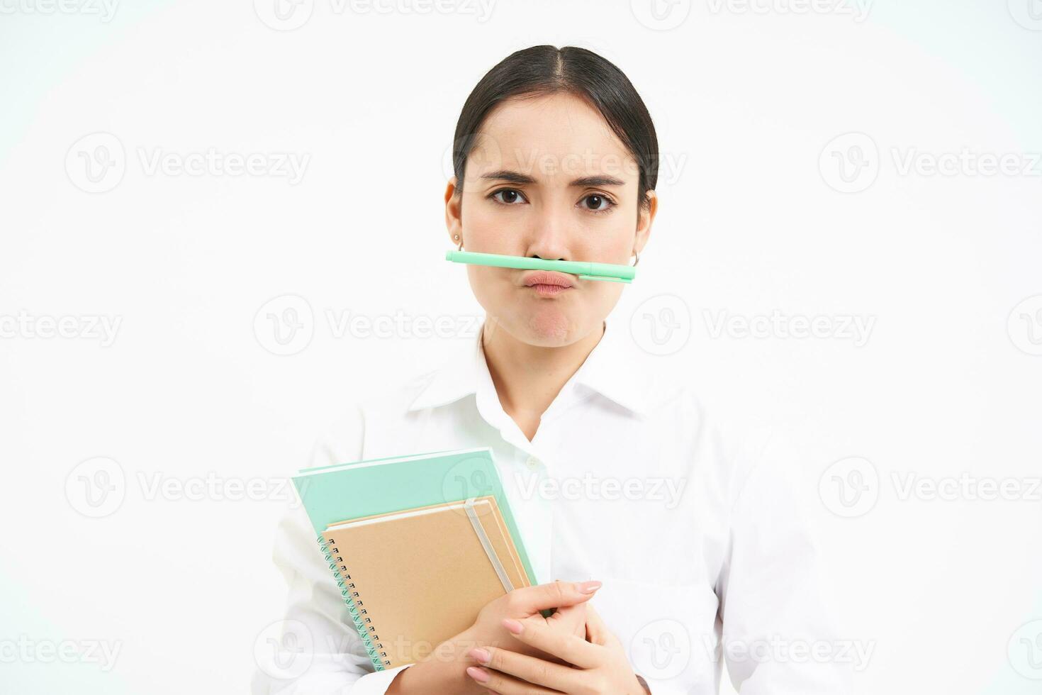 komisch Koreanisch Frau mit Notizbücher, hält Stift mit Lippen und Nase und macht Gesichter, steht Über Weiß Studio Hintergrund foto