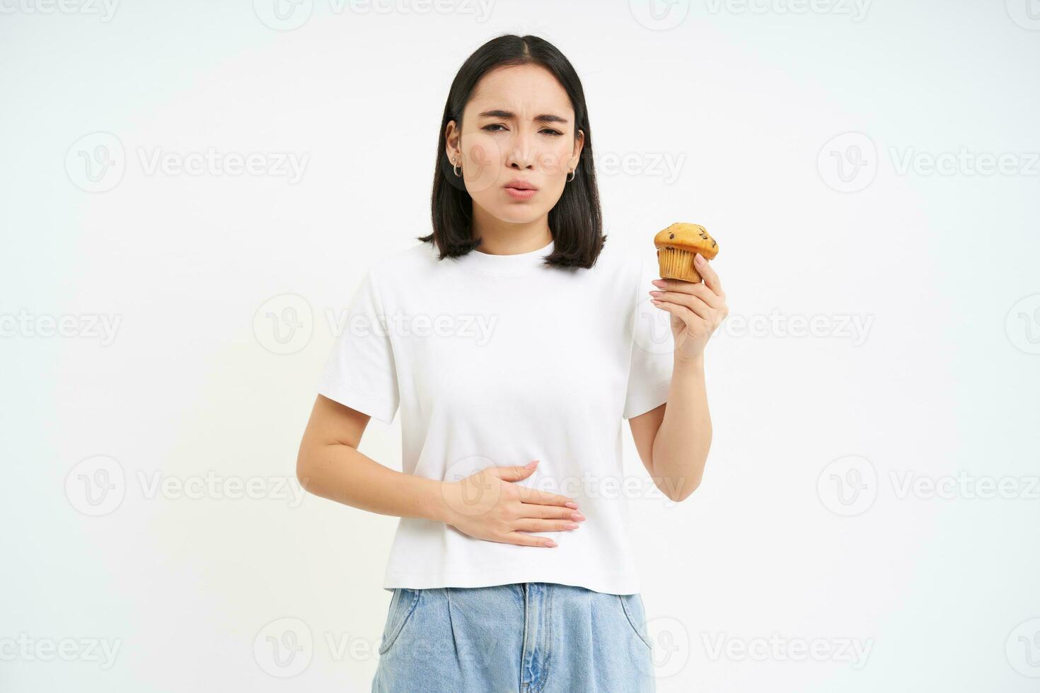 Porträt von Koreanisch Frau hat Bauch schmerzen, berühren Bauch, halten Cupcake, kippen Essen Gebäck, Weiß Studio Hintergrund foto