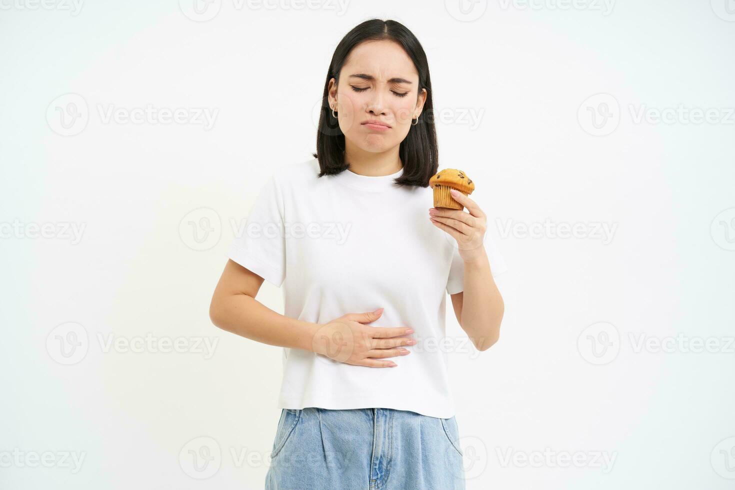 Porträt von Koreanisch Frau hat Bauch schmerzen, berühren Bauch, halten Cupcake, kippen Essen Gebäck, Weiß Studio Hintergrund foto