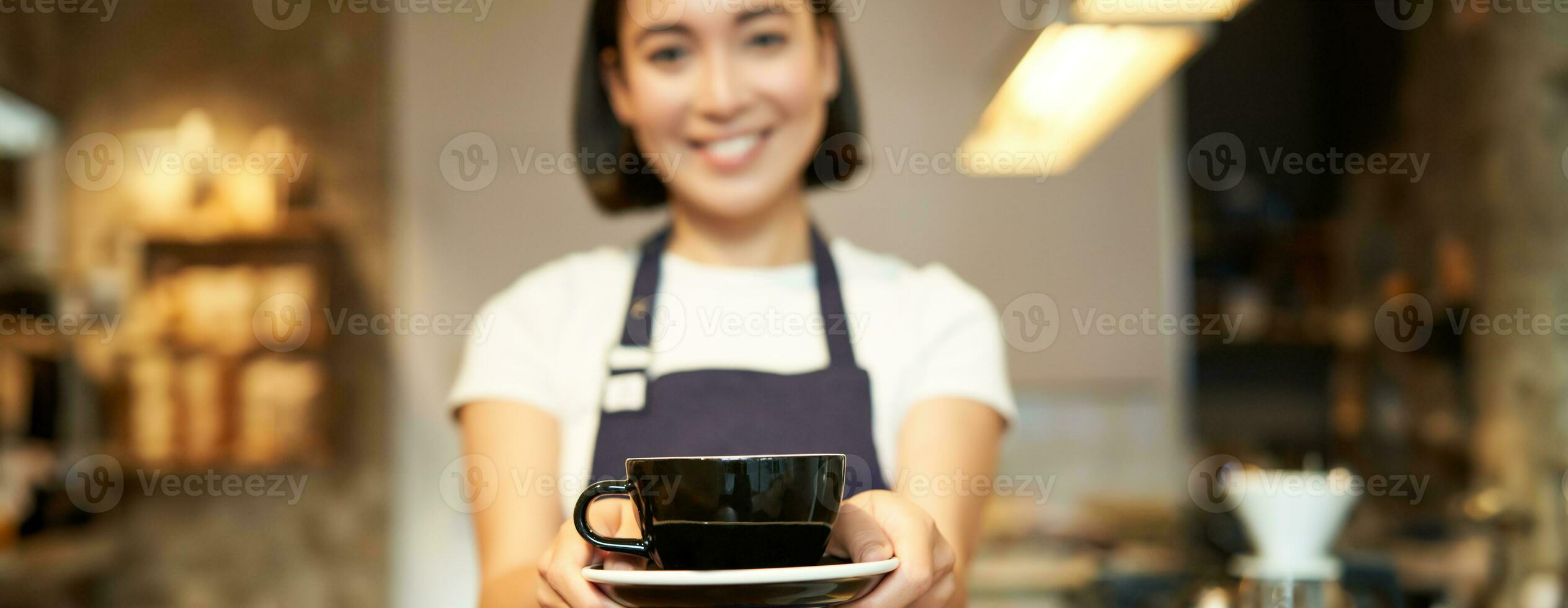 lächelnd asiatisch Mädchen Barista, Stehen im Schürze Uniform, geben Sie Tasse von Kaffee, gemacht ein trinken zum Klient im Cafe foto