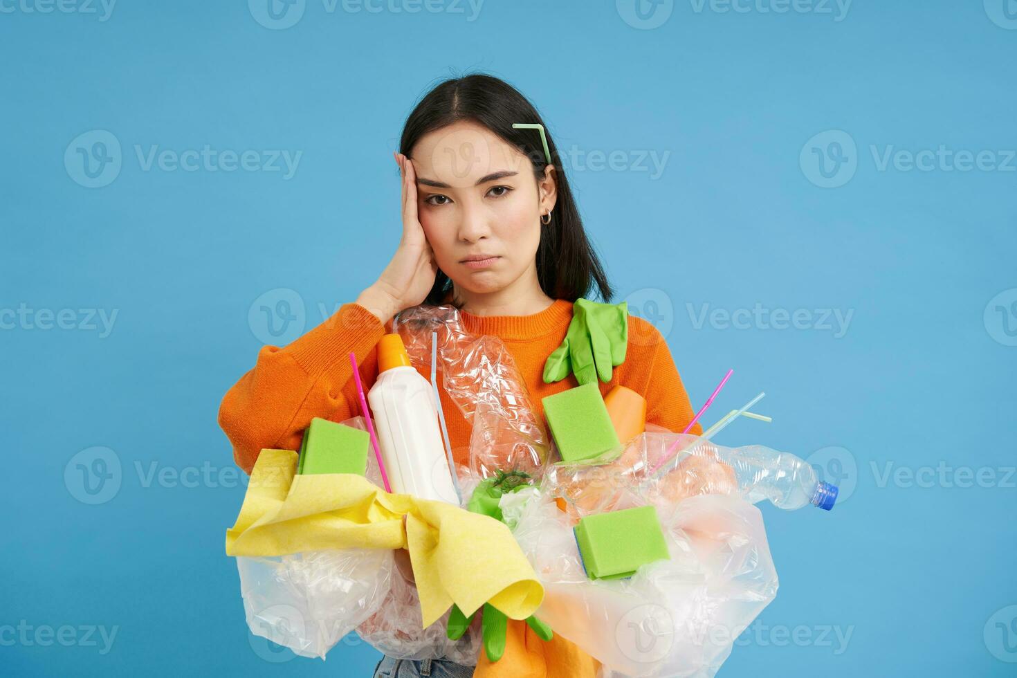 genervt jung Frau Reinigung Haus, halten Plastik Müll zum Recycling, müde von Sammeln Abfall, Stehen Über Blau Hintergrund foto