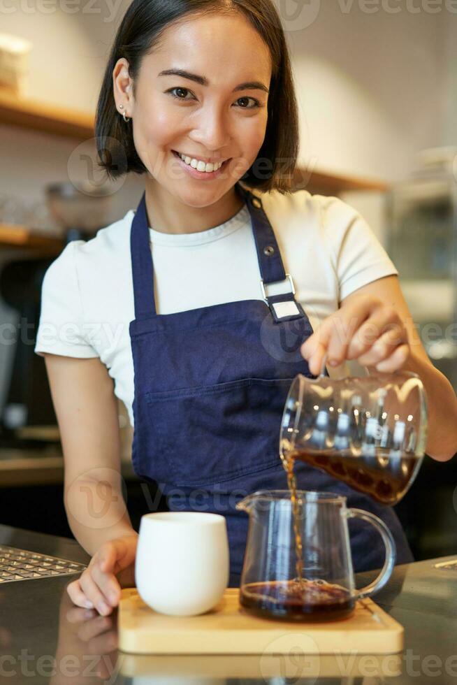 Vertikale Schuss von lächelnd süß Barista Mädchen, Gießen Filter Kaffee, Herstellung bestellen zum Cafe Klient, tragen Blau Schürze foto