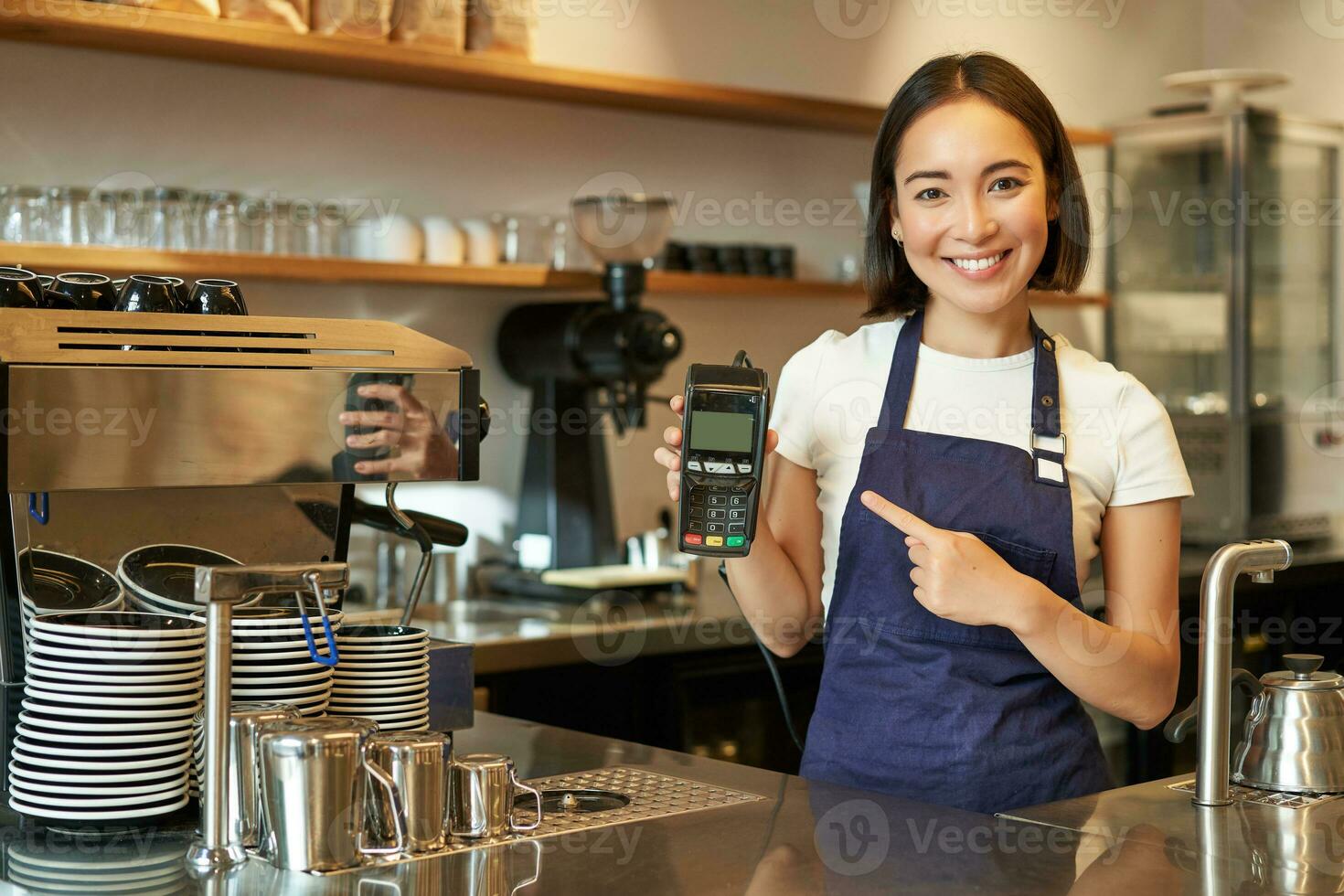 lächelnd asiatisch Mädchen Barista, Cafe Inhaber im Schürze, zeigen Karte Maschine, Zahlung Leser, nehmen kontaktlos Aufträge im ihr Kaffee Geschäft foto