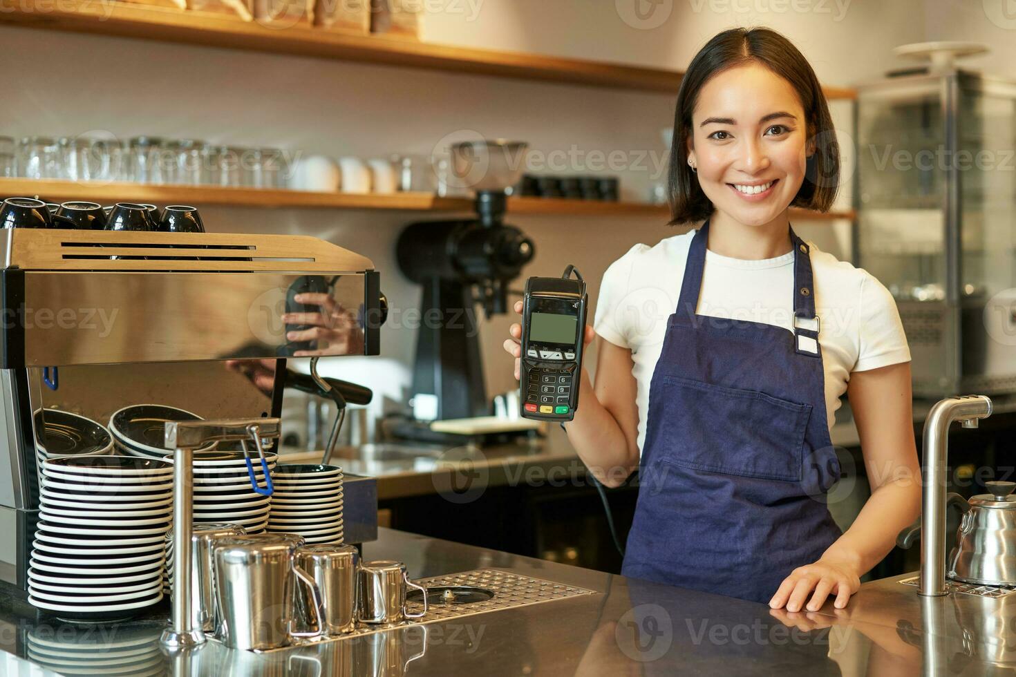 lächelnd asiatisch Mädchen Barista, Cafe Inhaber im Schürze, zeigen Karte Maschine, Zahlung Leser, nehmen kontaktlos Aufträge im ihr Kaffee Geschäft foto