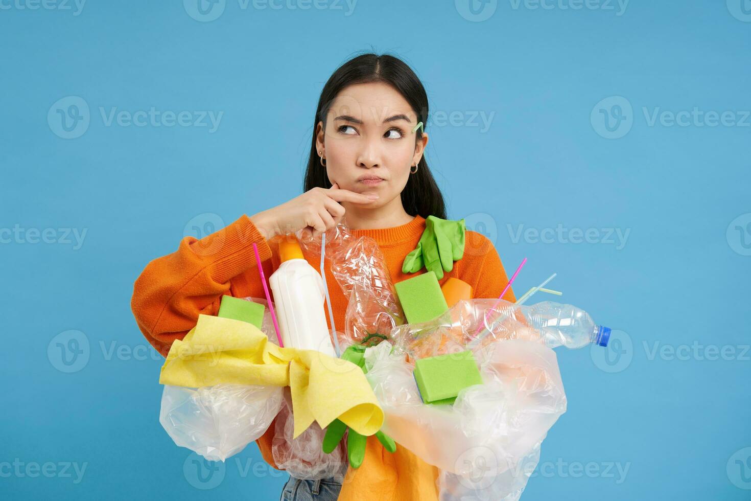 asiatisch Frau hält Plastik Abfall und macht Denken Gesicht, grübelt Wie zu recyceln Flaschen und Haushalt Artikel, Blau Hintergrund foto