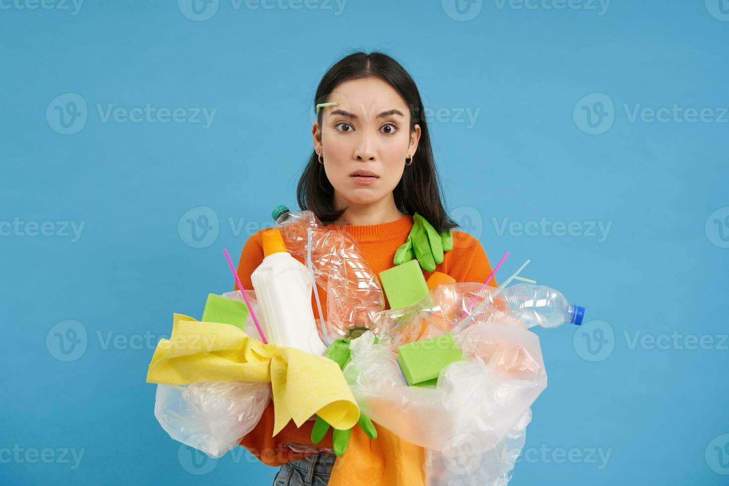 verwirrt asiatisch Mädchen nicht kennt Wie zu recyceln. Frau hält Plastik Müll mit schockiert Gesicht, Blau Hintergrund foto