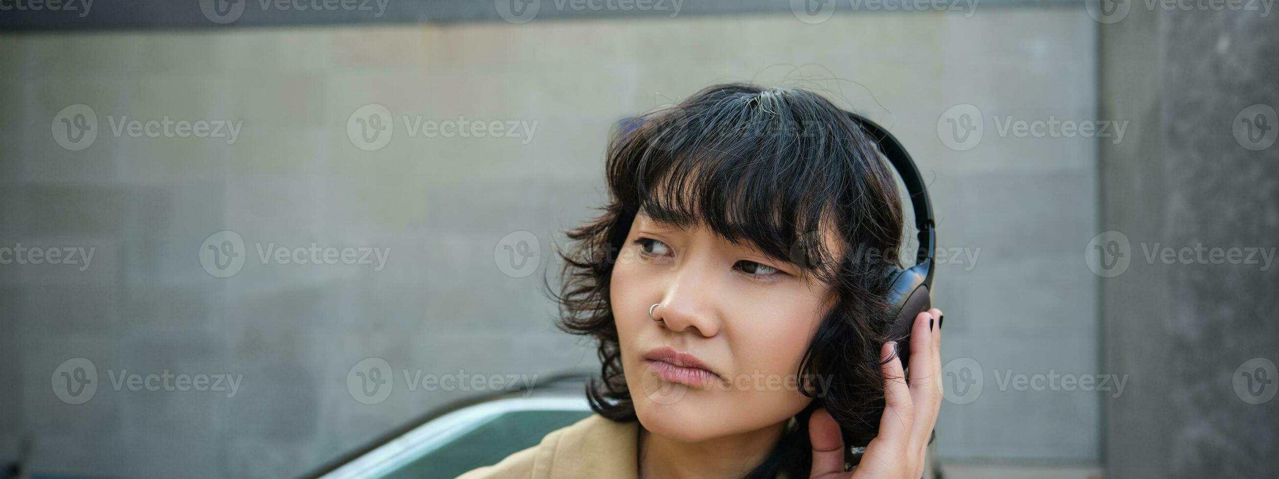 Porträt von kompliziert jung Koreanisch Mädchen im Kopfhörer, runzelt die Stirn und sieht aus enttäuscht während steht und wartet zum Öffentlichkeit Transport auf Straße foto