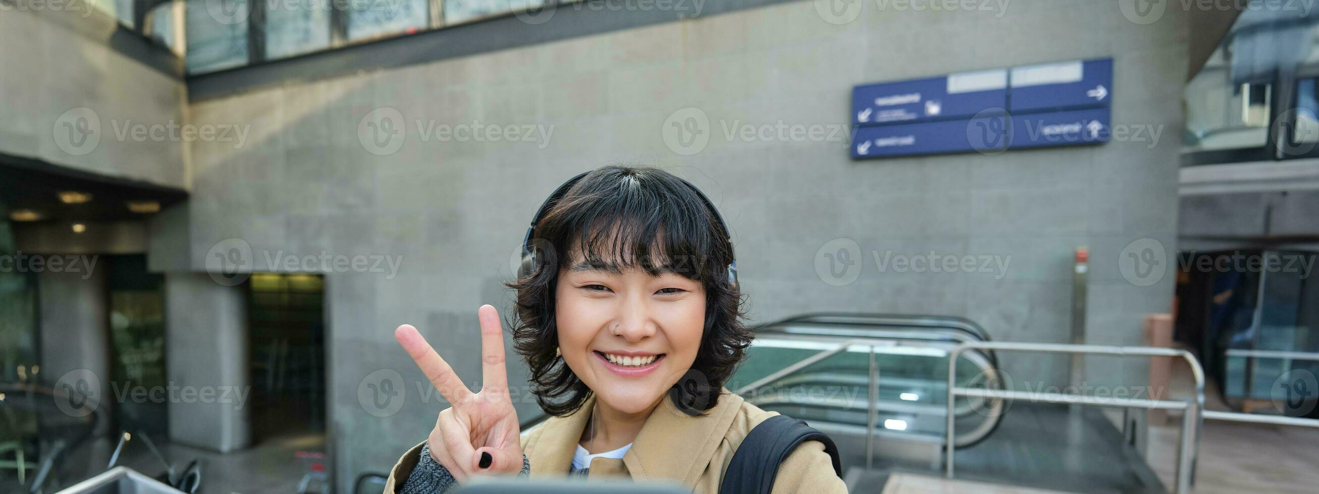 süß und stilvoll Koreanisch Mädchen, trägt Kopfhörer, nimmt Selfie auf Smartphone, Tourist Aufzeichnungen Video oder macht ein Foto, steht auf Straße foto
