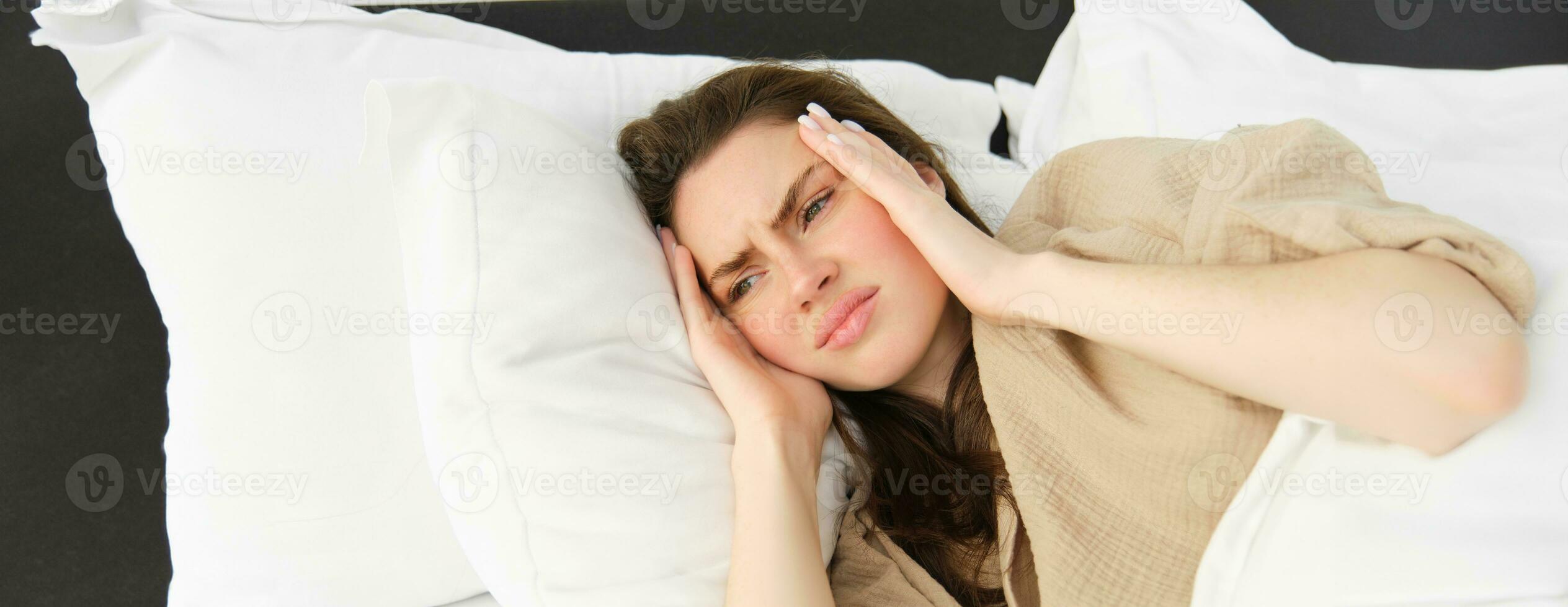 schließen oben Porträt von krank Frau Lügen im ihr Bett und Gefühl nicht wohl, berühren Kopf, hat Kopfschmerzen, suchen frustriert von Migräne, tragen Pyjama foto