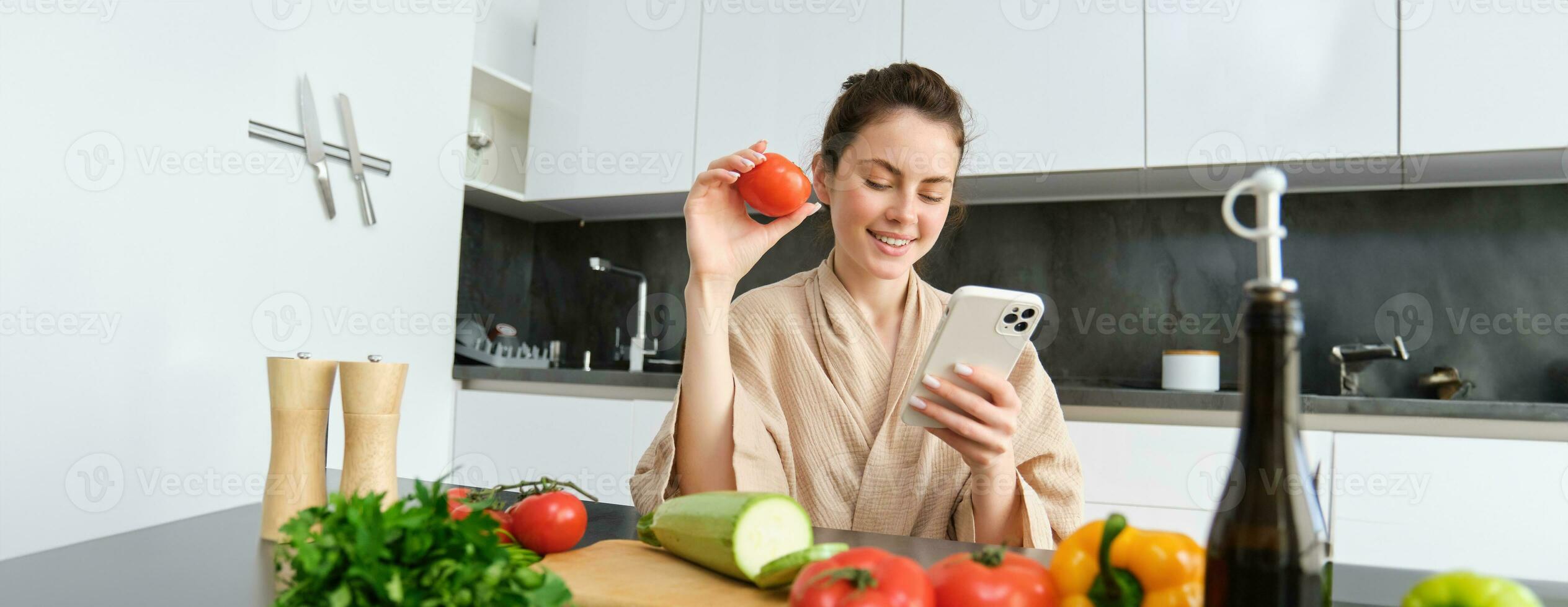 jung Frau Aufträge Lebensmittel auf Handy, Mobiltelefon App. Mädchen im Bademantel sitzt im das Küche mit Gemüse, suchen zum Rezept zu Koch Abendessen, mit Smartphone Anwendung foto