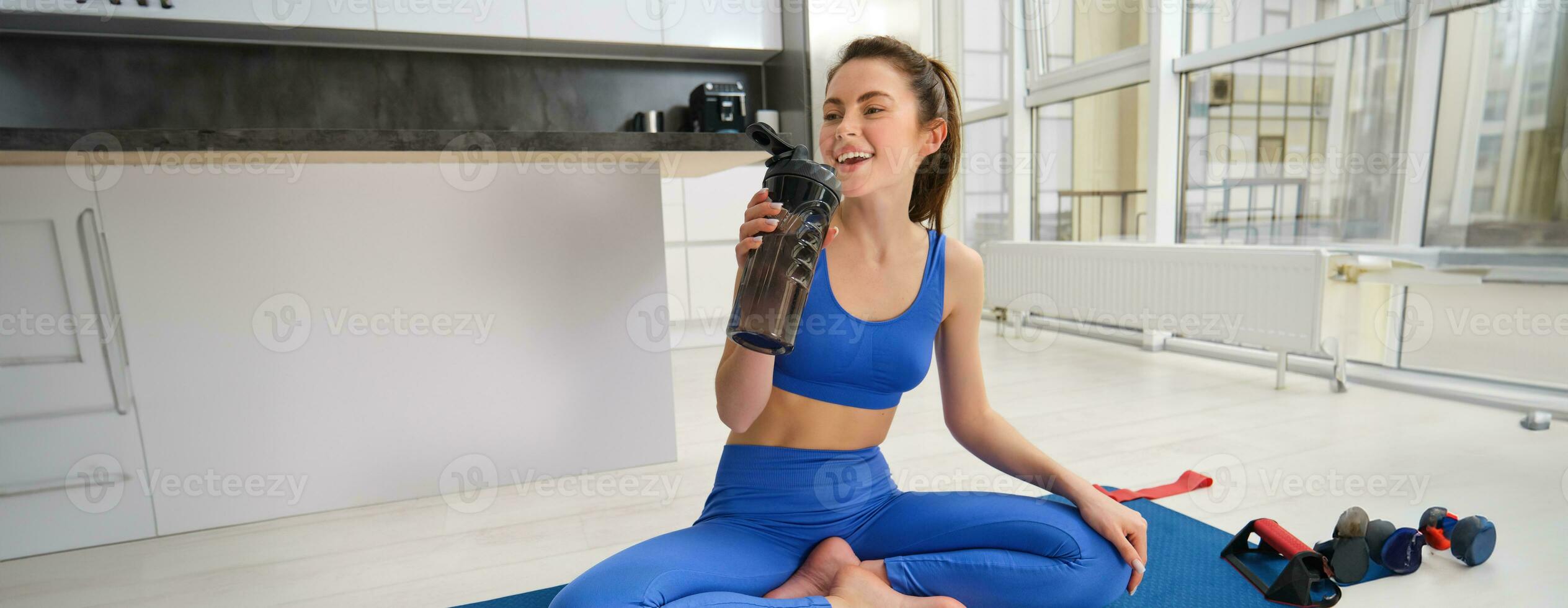 jung Frau Arbeiten aus von heim, Trinken Wasser während ihr Sport Ausbildung Session im Leben Zimmer, Sitzung auf Yoga Matte foto