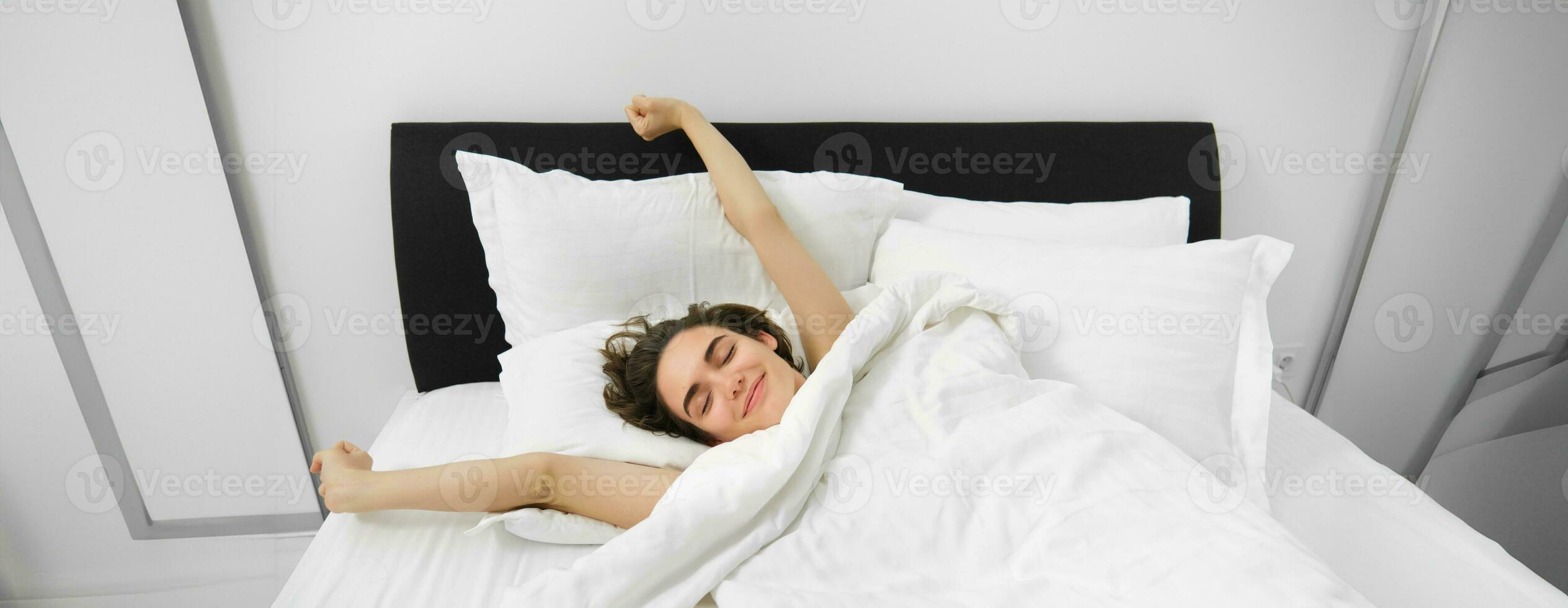 Porträt von Glücklich, zufrieden jung Frau, Dehnen ihr Waffen mit zufrieden lächeln, Lügen im Bett von Sanft Kopfkissen, aufwachen oben allein im Bett, hätten gut Nacht Schlaf foto