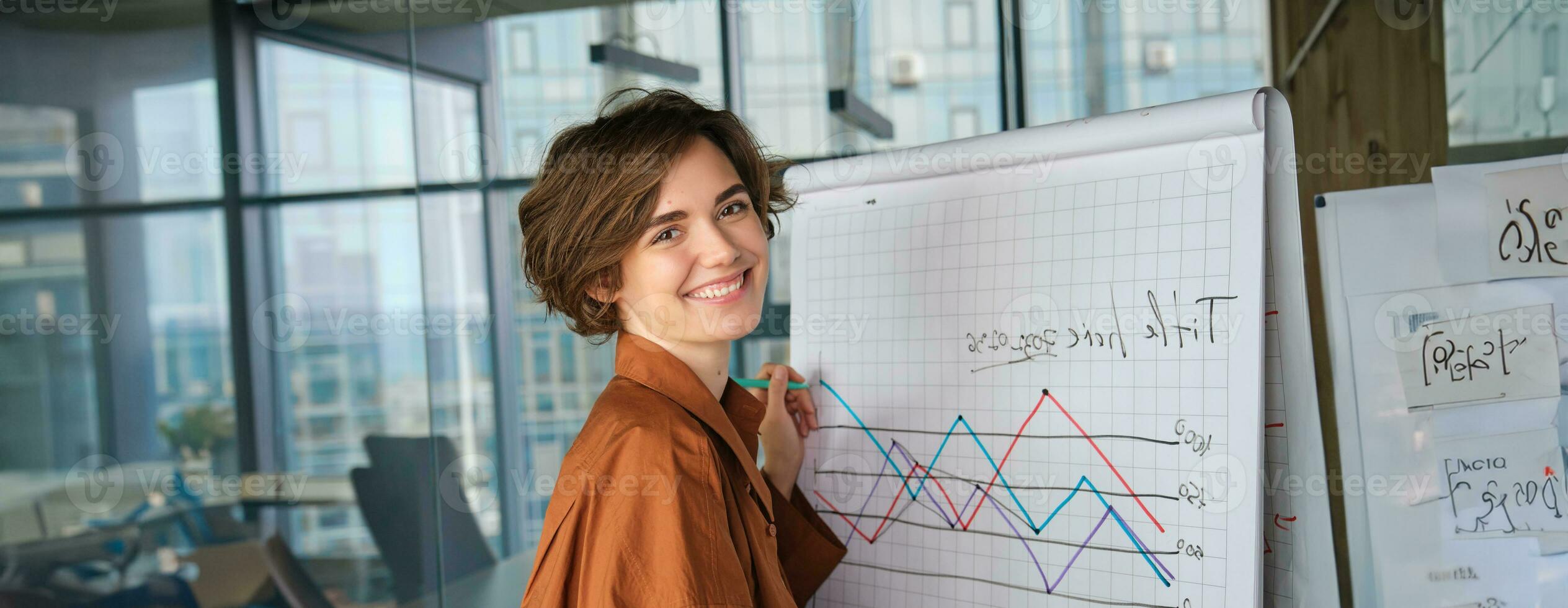 Porträt von jung Geschäftsfrau, Digital Nomade Stehen mit Marker in der Nähe von Tafel im Büro, Zeichnung ein Diagramm, geben Präsentation im Vorderseite von Team, lächelnd beim Kamera foto