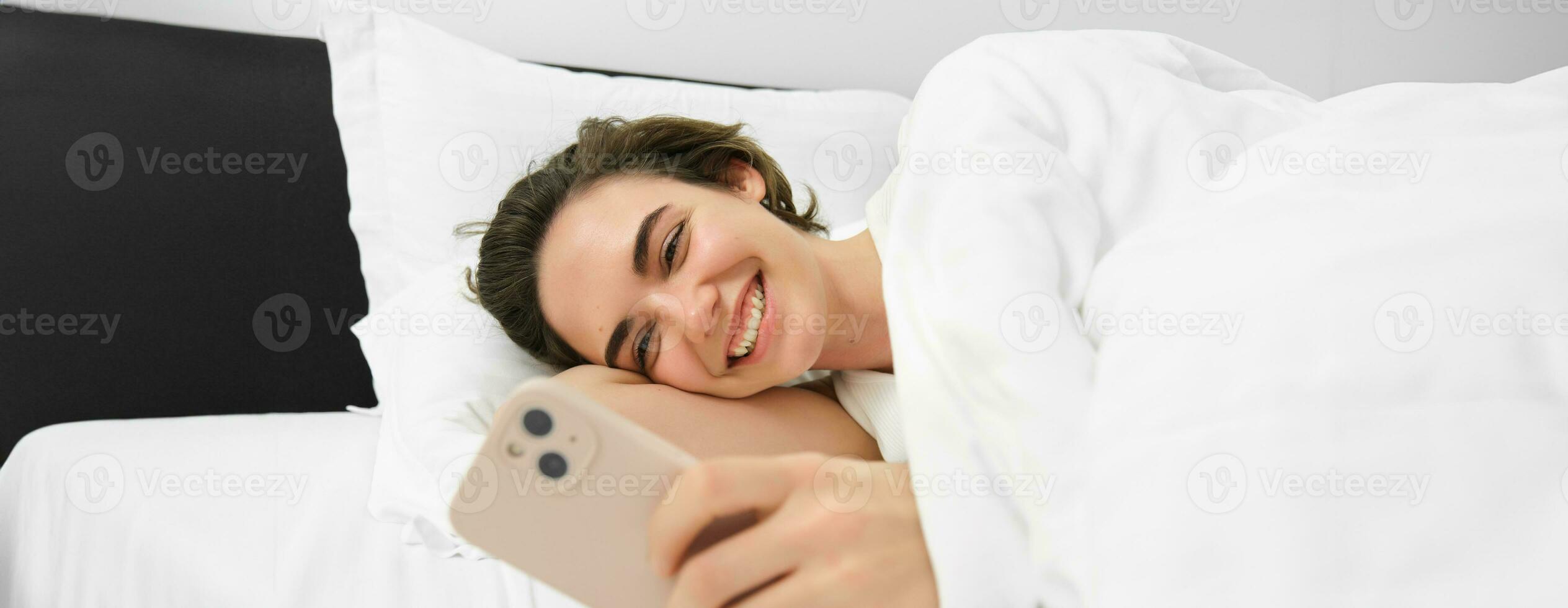 Bild von Frau Lügen im Bett mit Weiß Leinen- Blätter, suchen beim Smartphone und Lachen, Aufpassen komisch Video auf Handy, Mobiltelefon Telefon App foto