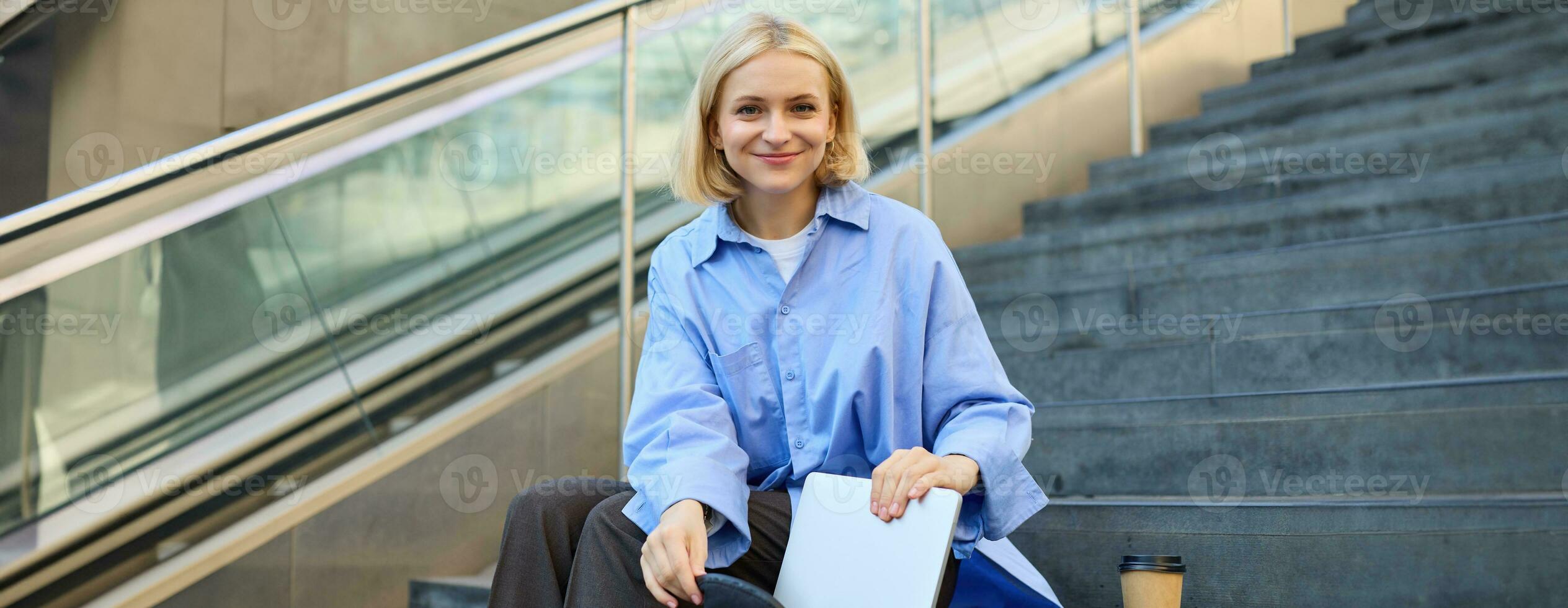 Porträt von schön lächelnd Frau, Schüler auf Treppe im Stadt, Sitzung und Trinken Kaffee, Verpackung Laptop im Rucksack, suchen glücklich beim Kamera foto