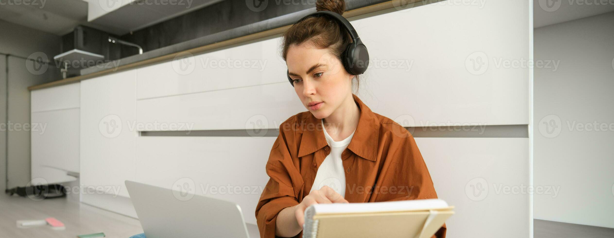 ernst aussehend Mädchen Schüler studieren beim heim, sitzt mit Laptop und Notizbuch auf Boden, tun Hausaufgaben Projekt auf Computer, hat online Kurs Lektion, trägt Kopfhörer foto