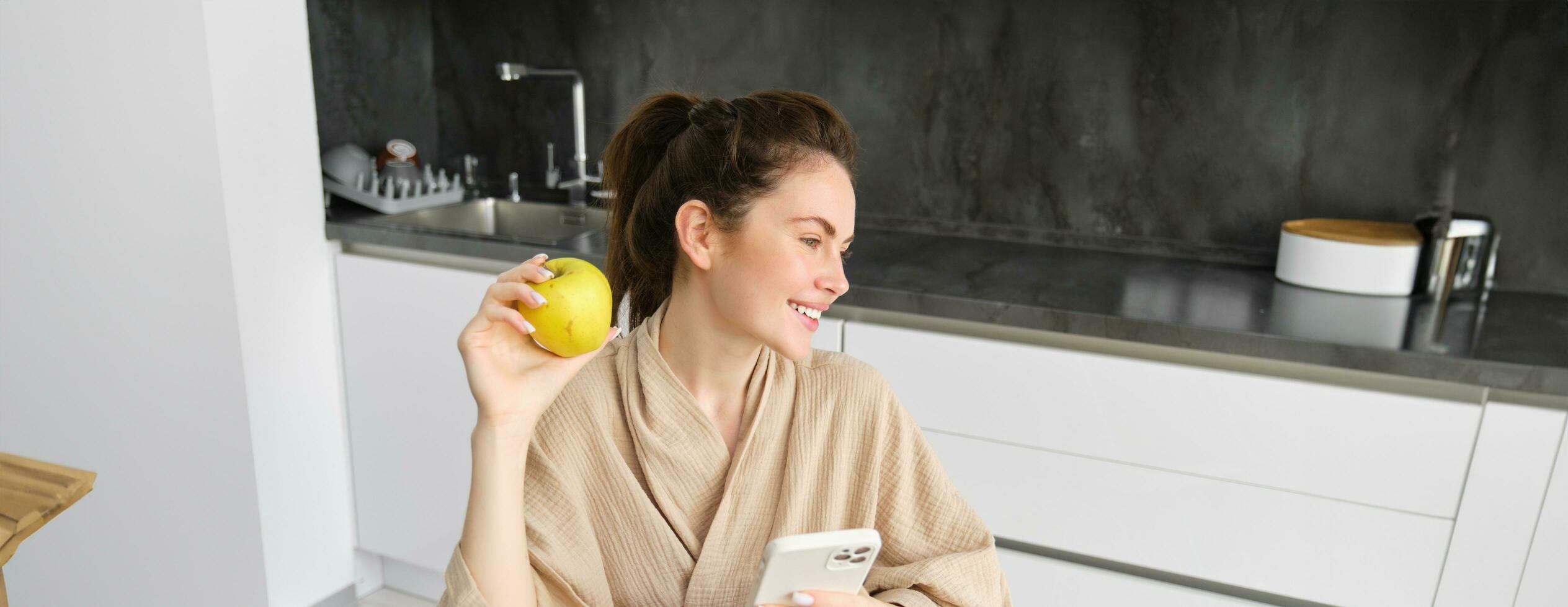 Bild von attraktiv jung Frau im Bademantel, sitzt im Küche, isst Grün Apfel und sieht aus beim Handy, Mobiltelefon Telefon, Verwendet Smartphone Anwendung, Aufträge Essen auf Anwendung foto