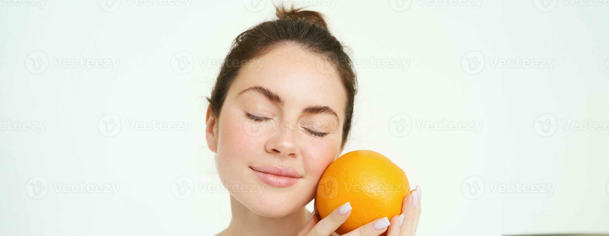 Porträt von jung Frau halten Orange in der Nähe von Gesicht ohne Schönheitsfehler, lächelnd, Konzept von Vitamine, Nahrung und organisch Gesichts- Behandlungen, Weiß Hintergrund foto